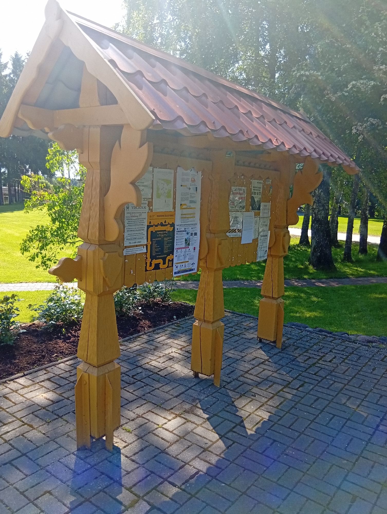 The image displays a large, elaborately carved wooden bulletin board with a tiled roof, situated outdoors on a paved area. The bulletin board is mounted on four substantial wooden posts, each intricately shaped and joined. Several notices and posters are affixed to the front of the bulletin board. The setting appears to be a park or public space, with lush green grass and trees visible in the background. The sunlight casts distinct shadows on the paved ground, indicating it is daytime. The overall impression is one of a rustic, well-maintained public information display in a natural environment. The image does not contain any people or discernible actions.