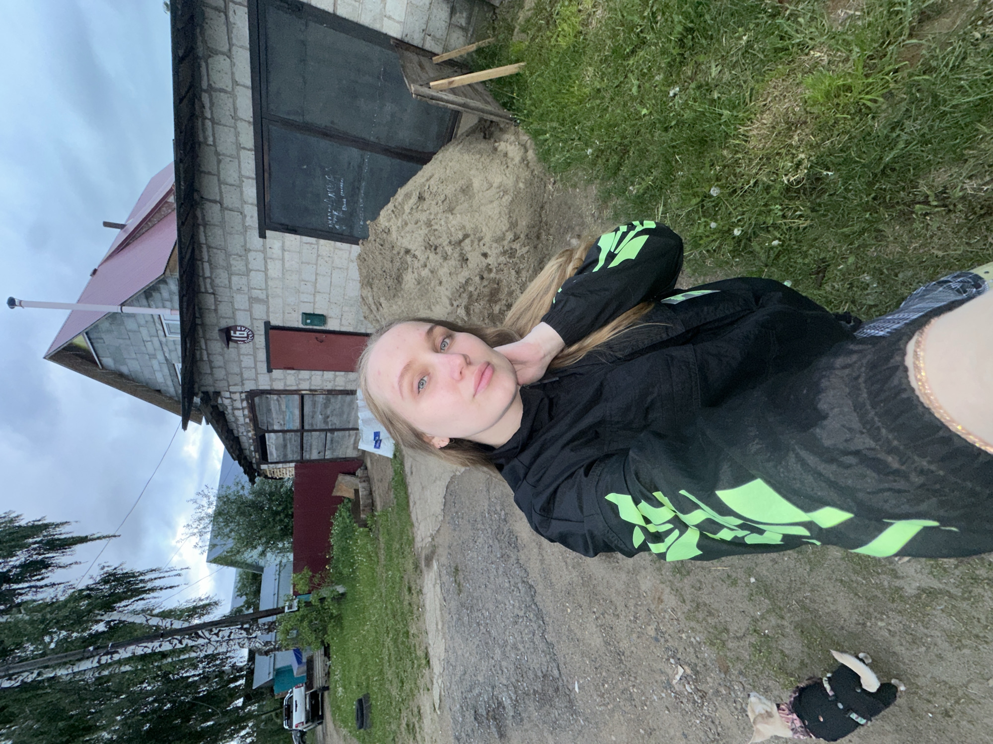 An outdoor, eye-level selfie captures a young adult female on a dirt path in Chaikovsky, Russia, under an overcast sky. The female, positioned center-left, has long blonde hair and light-colored eyes, and wears a black jacket with neon green graphic designs on the sleeves. Her right hand touches her neck. To her lower left, a small, light-colored dog in a black garment faces away from the camera.
The immediate foreground consists of a dirt and gravel path. To the right, a grassy area with sparse weeds extends towards a large pile of light brown sand. Leaning against the sand pile are several wooden planks. Behind the sand and the female, a single-story building constructed of light-colored cinder blocks features a red door with a green mailbox to its right. Above the door, a white circular sign with black text reads "16 СТАБИЛЬНАЯ". To the right of the red door, two large, dark-colored panels, possibly garage doors, are visible, one bearing white scribbled text.
Further back, behind the cinder block building, a two-story house with exposed cinder block walls and a red corrugated metal roof stands, with a white chimney pipe extending skyward. To the far left, several tall birch trees with prominent white bark and drooping green leaves frame a dirt road. A white pickup truck is partially visible on this road in the background. A dark red metal fence separates properties in the mid-ground. Other distant buildings with light-colored roofs are visible through the trees. A utility pole is situated behind the birch trees. A discarded tire rests on the ground near the red fence on the left.