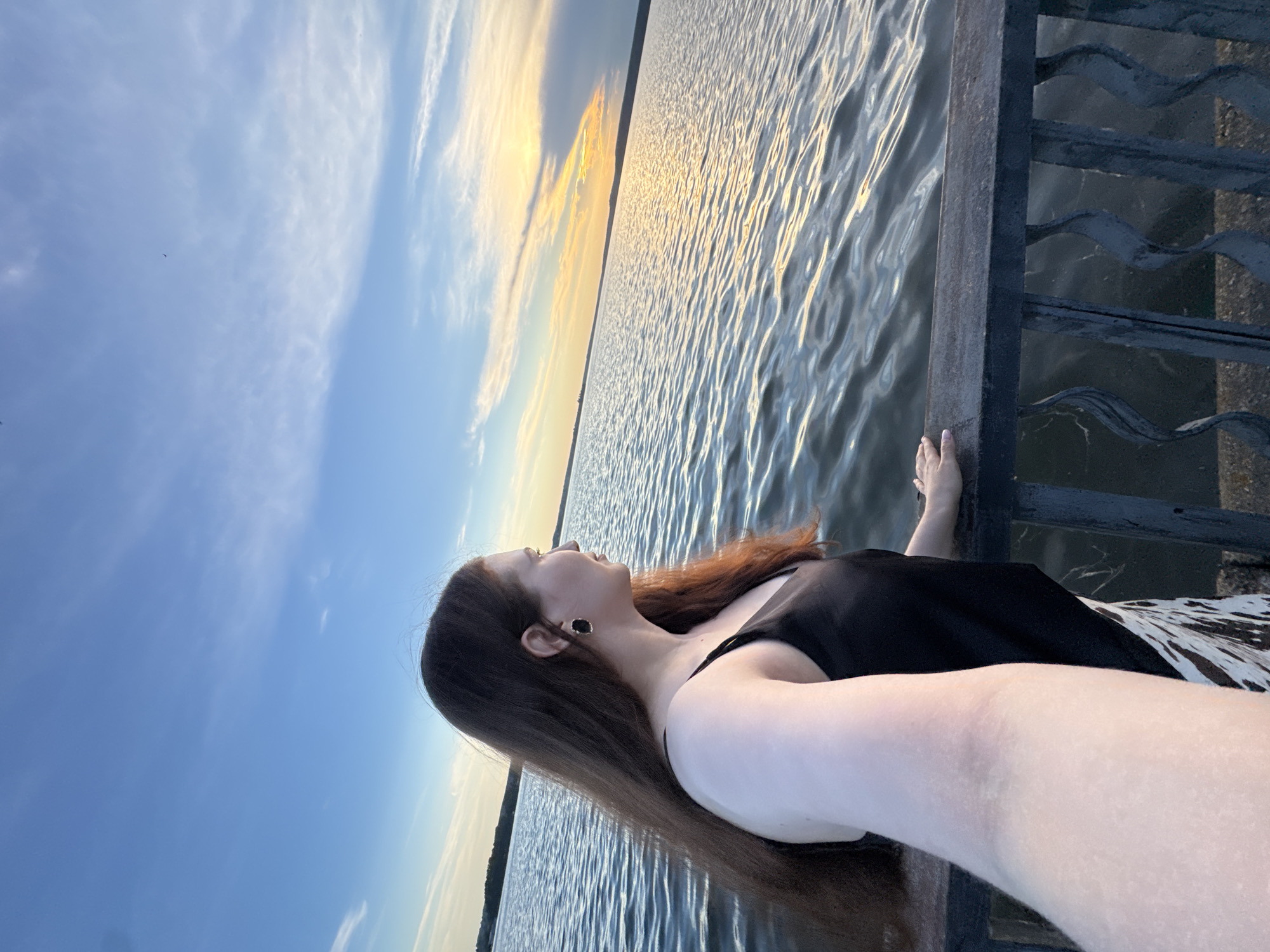 A young woman with long brown hair and black earrings stands on a pier, looking out at a body of water during sunset. The sky is a mix of blue and orange, with wispy clouds. The water is rippled, reflecting the colors of the sky. The woman is wearing a black tank top and a patterned skirt. The pier is made of dark metal and wood. The location is Votkinsk, Russia.