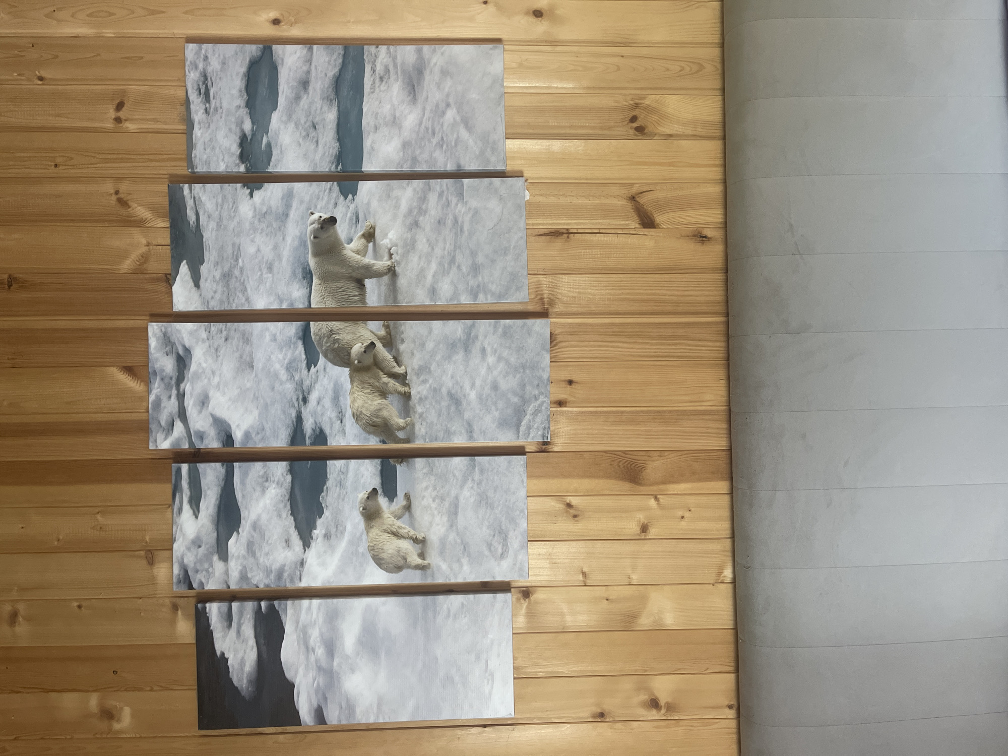 The image displays a five-panel canvas print of polar bears on an icy landscape, mounted on a wooden paneled wall. The print features a mother polar bear walking with two cubs on ice floes with patches of dark water. The overall scene depicts an Arctic environment. The location is stated to be in Bukhovoye, Russia. Below the artwork, a light gray upholstered headboard is visible.