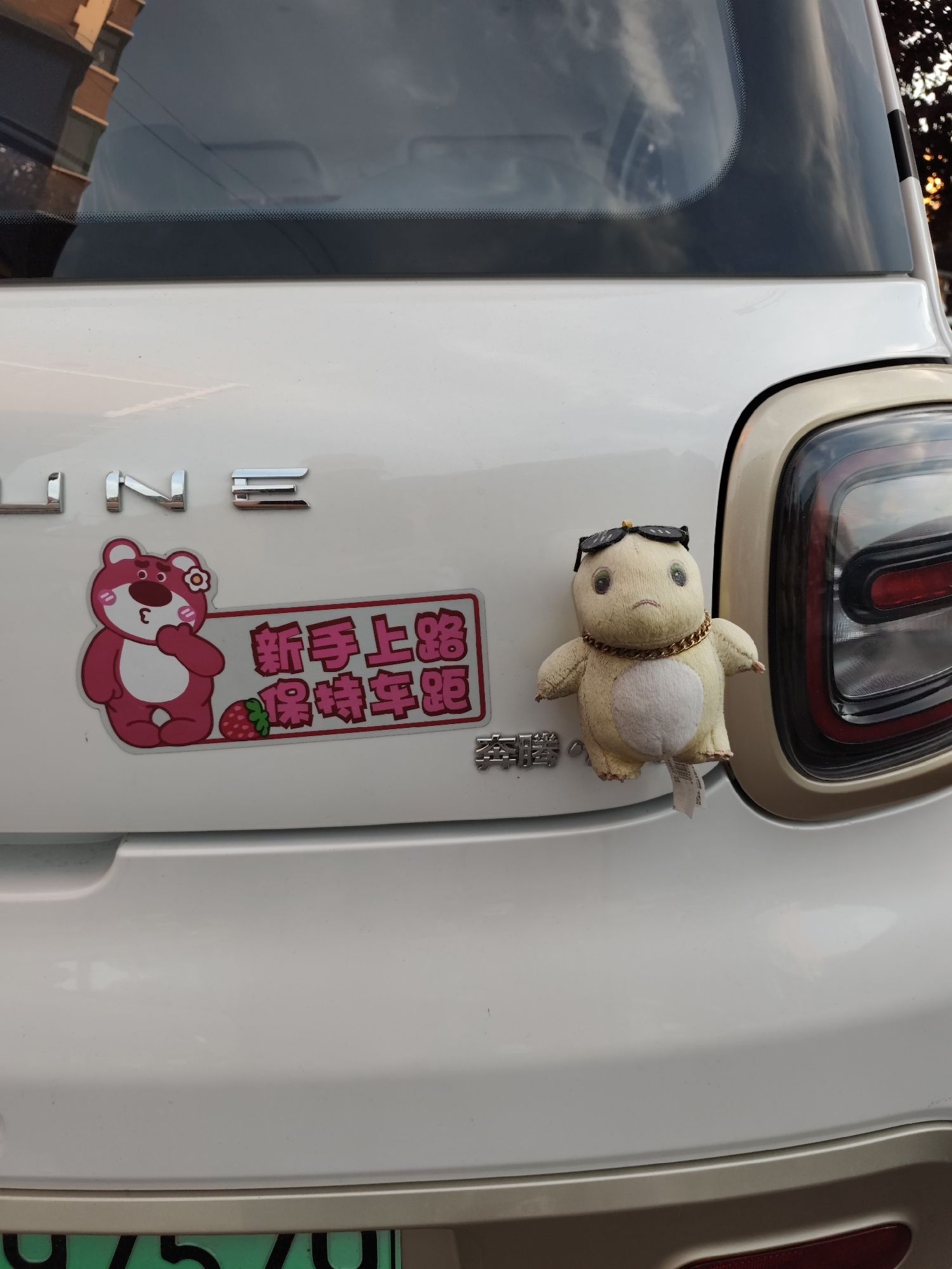 A white car is parked outdoors, likely in a city like Lindian, China, as evidenced by the presence of a Chinese license plate. The car's rear is visible, featuring a taillight and a bumper. Attached to the bumper are two decorative items: a pink sticker of a cartoon bear with a strawberry, and a small, fuzzy stuffed toy wearing sunglasses and a gold chain. The sticker reads "新手上路保持车距" in Chinese, which translates to "Novice on the road, maintain distance." The plush toy is positioned to the right of the sticker. The car appears to be in good condition, and the scene suggests a typical urban or suburban setting.