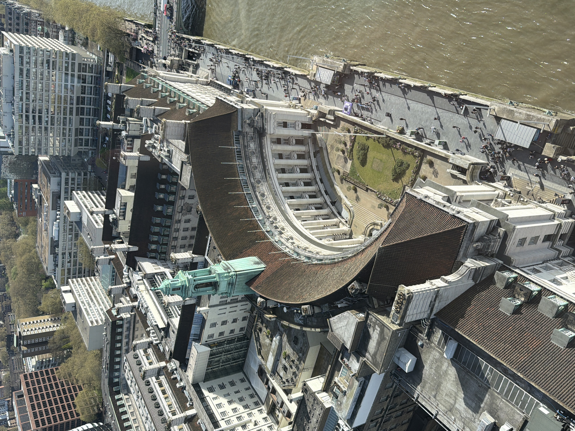 This is an aerial view of a section of London, United Kingdom, taken looking more or less straight down. In the foreground, rooftops of buildings in various architectural styles dominate the view. A prominent building with a curved facade, covered with a brown tiled roof, is visible, running alongside a plaza with a small garden, with the River Thames in the background. Along the riverbank, people are seen walking along the embankment. To the left, there is a modern building with a grid-like facade. A row of trees can be observed along the street, near the buildings. The water in the river appears murky.