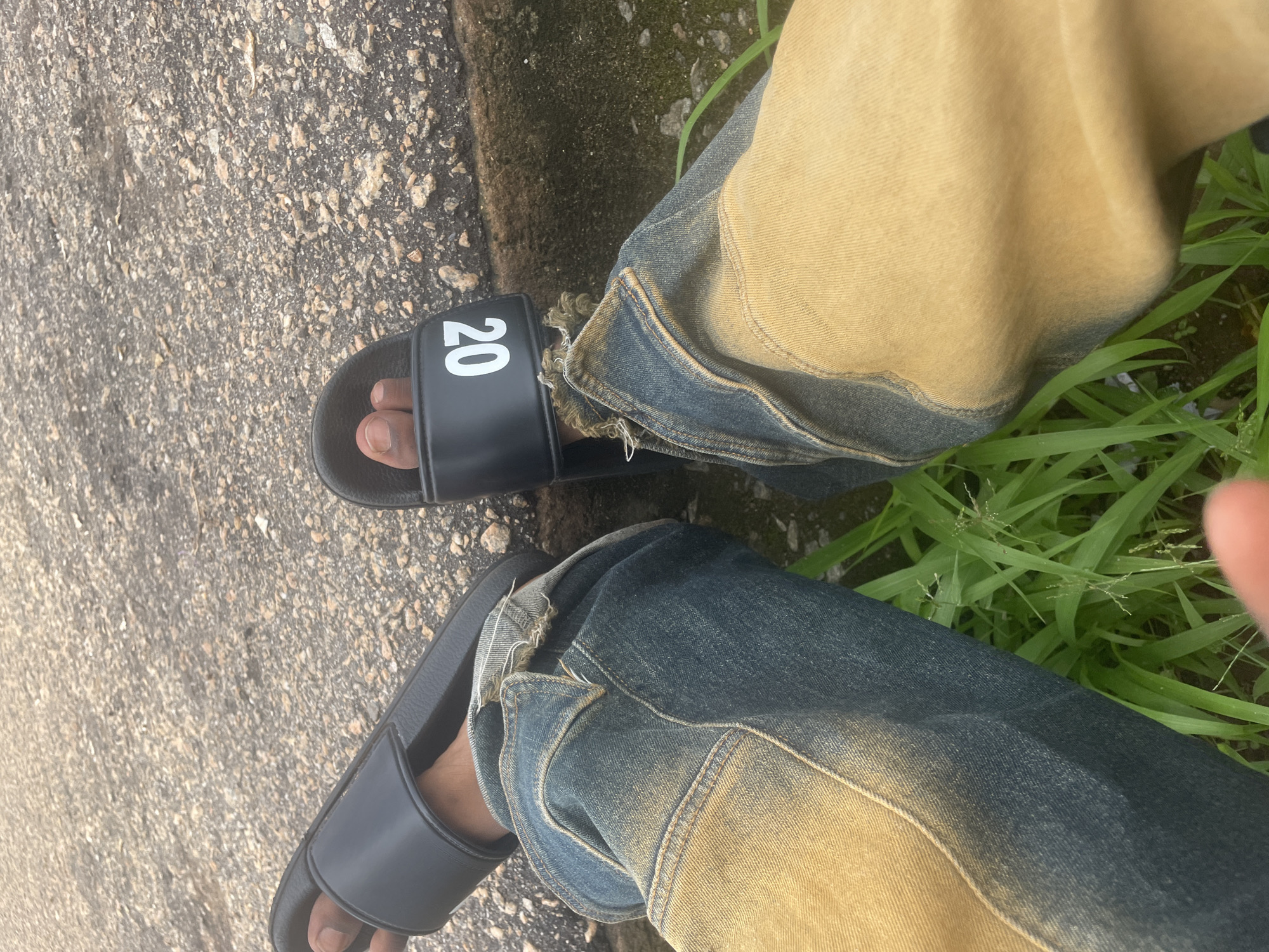 The image shows a close-up view of a person's legs and feet, from the knees down, wearing denim jeans and black slide sandals. The jeans are a faded blue with a yellowish-brown wash, and appear to be distressed with frayed edges near the ankles. One of the sandals has the number "20" printed in white on the strap. The person is sitting or resting on a rough, grey, concrete-like surface, possibly a pathway or roadside, with a thin strip of green grass visible to the right. The lighting suggests it could be daytime, with diffused light, and the overall scene is casual. The location context provided is Lokoja, Nigeria, but there are no specific visual cues within the image to confirm this. There are no other visible subjects, and the focus is entirely on the clothing and the immediate surroundings.