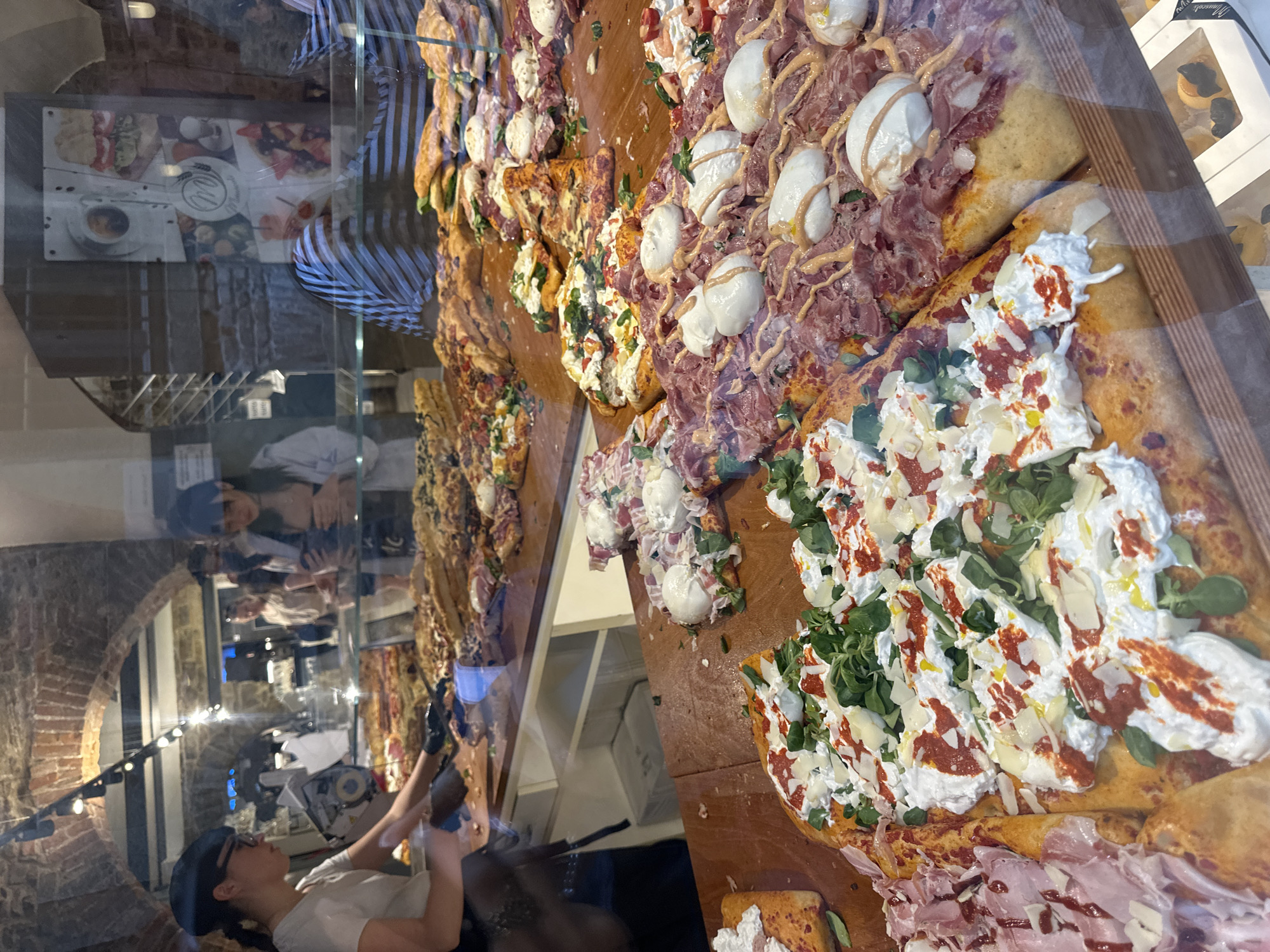 A display of various rectangular pizzas is presented at a shop in Bergamo, Italy. The pizzas feature a variety of toppings, including mozzarella, prosciutto, vegetables, and sauces. A person wearing a hat and t-shirt is visible in the background, seemingly preparing or serving the pizzas. The pizzas are displayed on a wooden surface behind a glass enclosure, and the reflection on the glass shows architectural details such as brick arches and interior lighting.