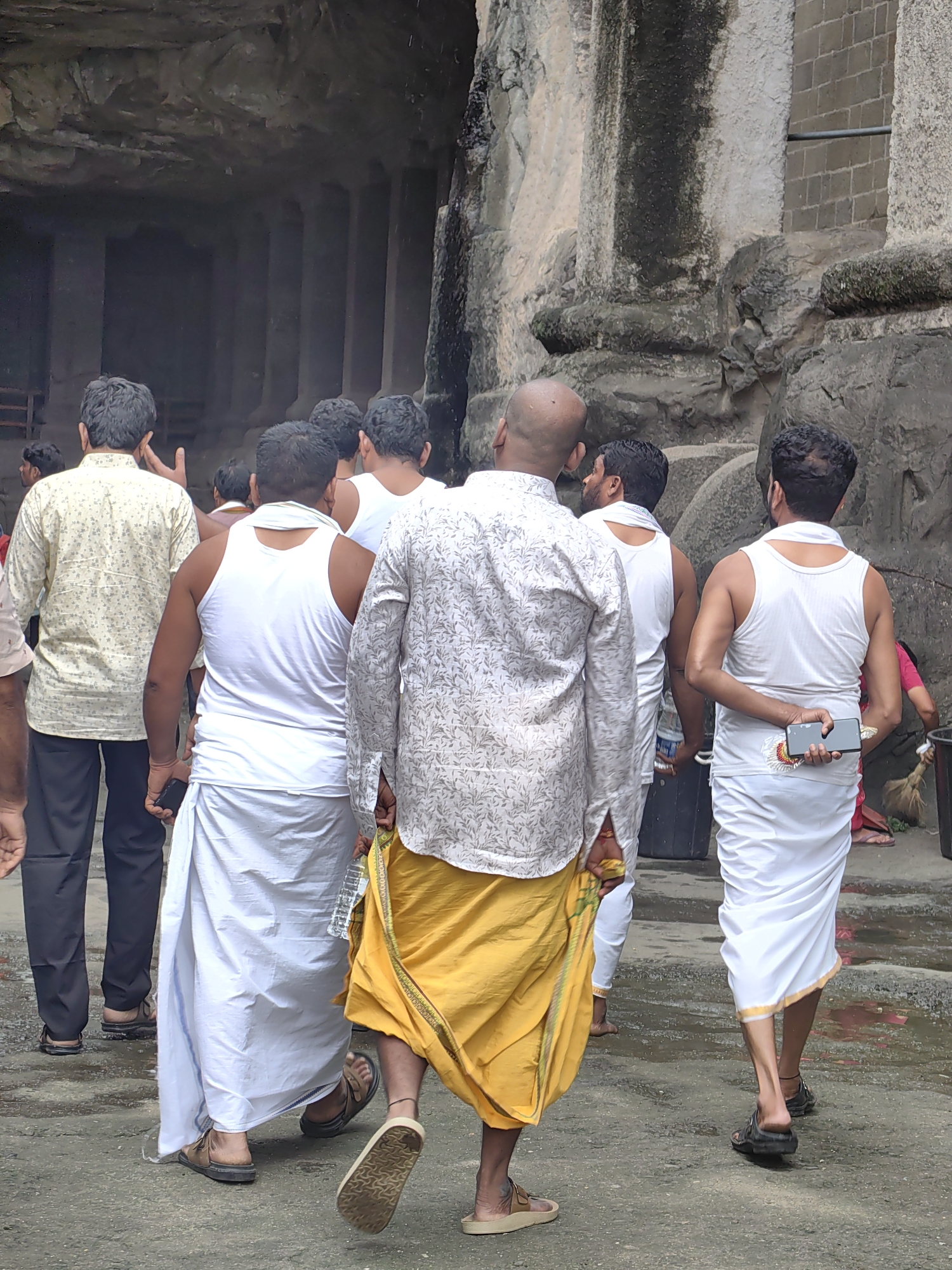 A group of men, some wearing traditional Indian attire of white dhotis and vests, and others in shirts and pants, walk away from the camera. The scene appears to be at the Ellora Caves, a UNESCO World Heritage site in Maharashtra, India, characterized by ancient rock-cut architecture. The men are walking on a wet, paved surface, possibly indicating recent rain or damp conditions. The background shows the imposing, textured stone walls of the caves. One man in the foreground is wearing a patterned white shirt and a bright yellow dhoti, and another man behind him is in a white vest and a white dhoti. Several other men are visible behind them, also dressed in similar traditional clothing. The overall atmosphere is one of a tourist or pilgrimage visit to a historical and religious site.
