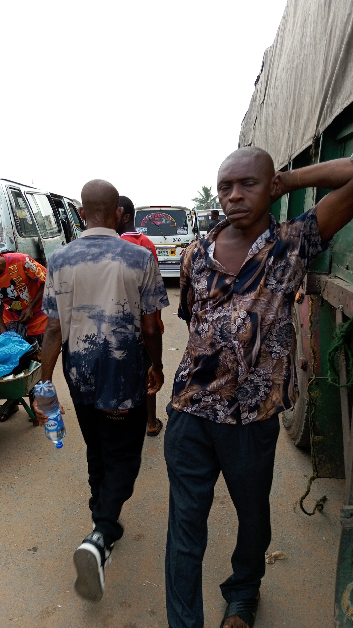 The image depicts an outdoor scene in what appears to be a busy area of Onitsha, Nigeria. A man in a dark, patterned short-sleeved shirt and dark pants stands with his arm resting on the side of a large truck. He is looking directly at the camera. Another man walks away from the camera, wearing a shirt with a landscape design and dark pants, carrying a bottle of water. Several vehicles are visible in the background, including a white van with the license plate "UT 9237 80" visible. The setting appears to be a roadside with other individuals and objects in the background, suggesting a commercial area.