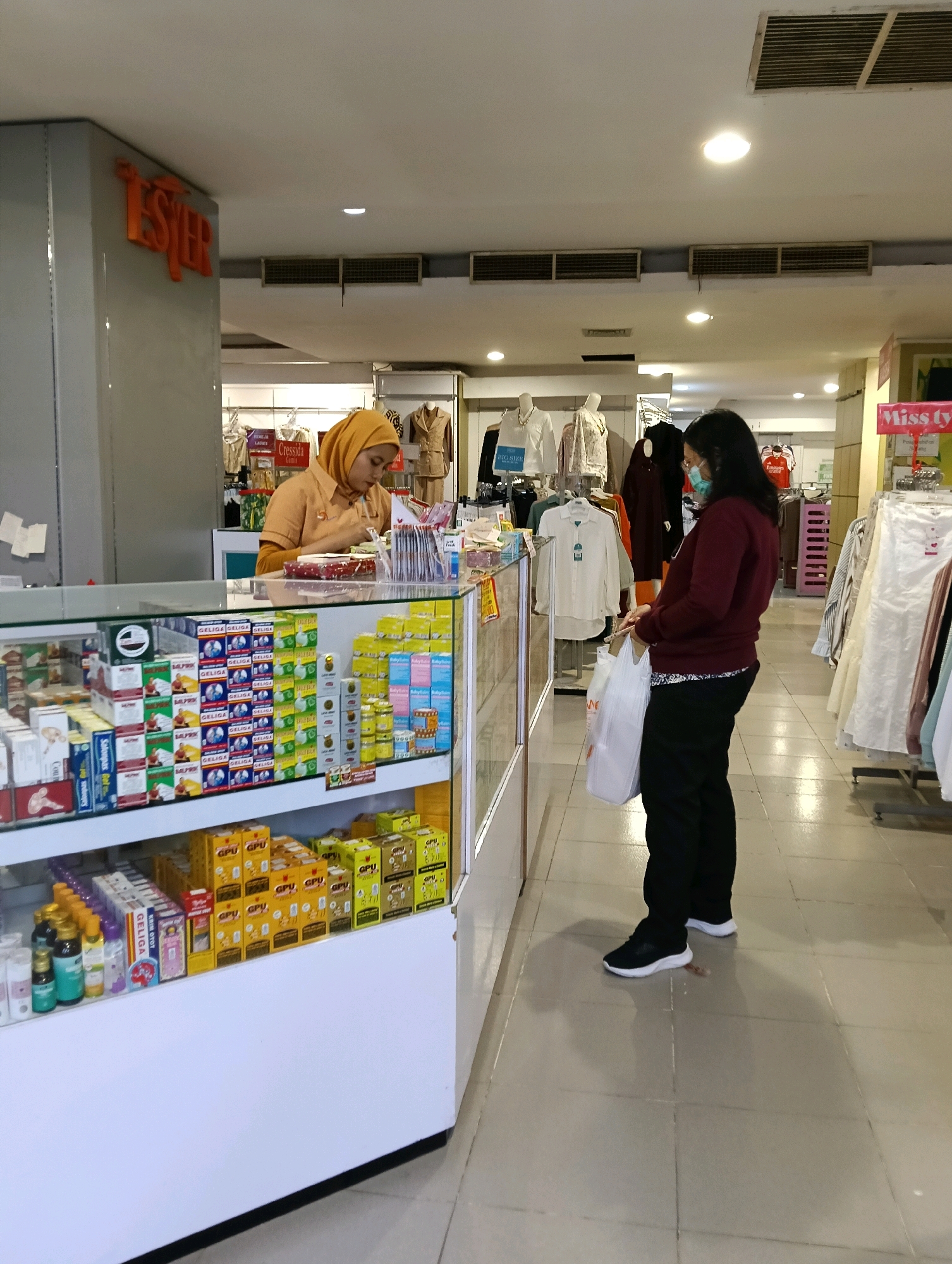In Jaten, Indonesia, inside a store, a person wearing a hijab, dressed in an orange uniform, stands behind a counter, interacting with a customer. The customer, a woman wearing a maroon sweater and black pants, also wears a light blue face mask and holds two white plastic bags. The counter is filled with various products, including boxes labeled "GELIGA" and "GPU," as well as bottles of liquid. In the background, racks of clothing are displayed, with mannequins wearing white shirts. A sign with orange lettering, reading "TESTER," is visible on the left side of the image. The store appears to be well-lit with overhead lighting.