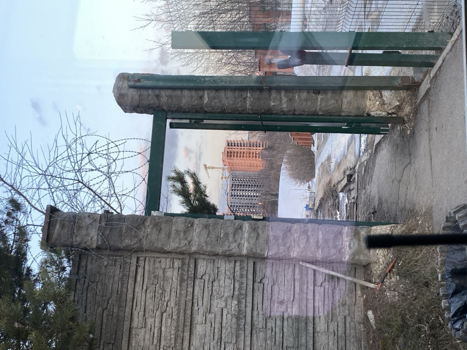 This image, located in Марфино, Russia, depicts a scene through a partially open green metal gate and an adjacent concrete wall. The foreground shows a black cylindrical object in the lower center, and part of a wooden planter containing a black trash bag in the lower left. Reflections are visible on the surface from which the photograph was taken, suggesting a glass barrier.
On the left, a textured grey concrete wall stands, adjoined by a substantial concrete gate post topped with coils of barbed wire. A green pine branch extends from above the wall into the frame. Leaning against the wall near its base is a tool resembling a shovel or rake with a red handle.
The open gate, constructed of green metal bars and supported by a second concrete post, reveals a downward-sloping, cracked asphalt path covered in patches of mud, dirt, and melting snow. Bare deciduous trees and shrubs line the path beyond the gate. To the far right, a segment of a metal wire fence is visible.
In the mid-distance, a landscape of sparse winter vegetation and patches of snow leads to a construction site. Two multi-story buildings are under construction: one grey and incomplete, and another reddish-orange brick structure, illuminated by warm, possibly sunset light. A yellow tower crane is positioned between these buildings. The sky above is partially cloudy, displaying shades of blue, pink, and orange. No individuals are present in the scene.