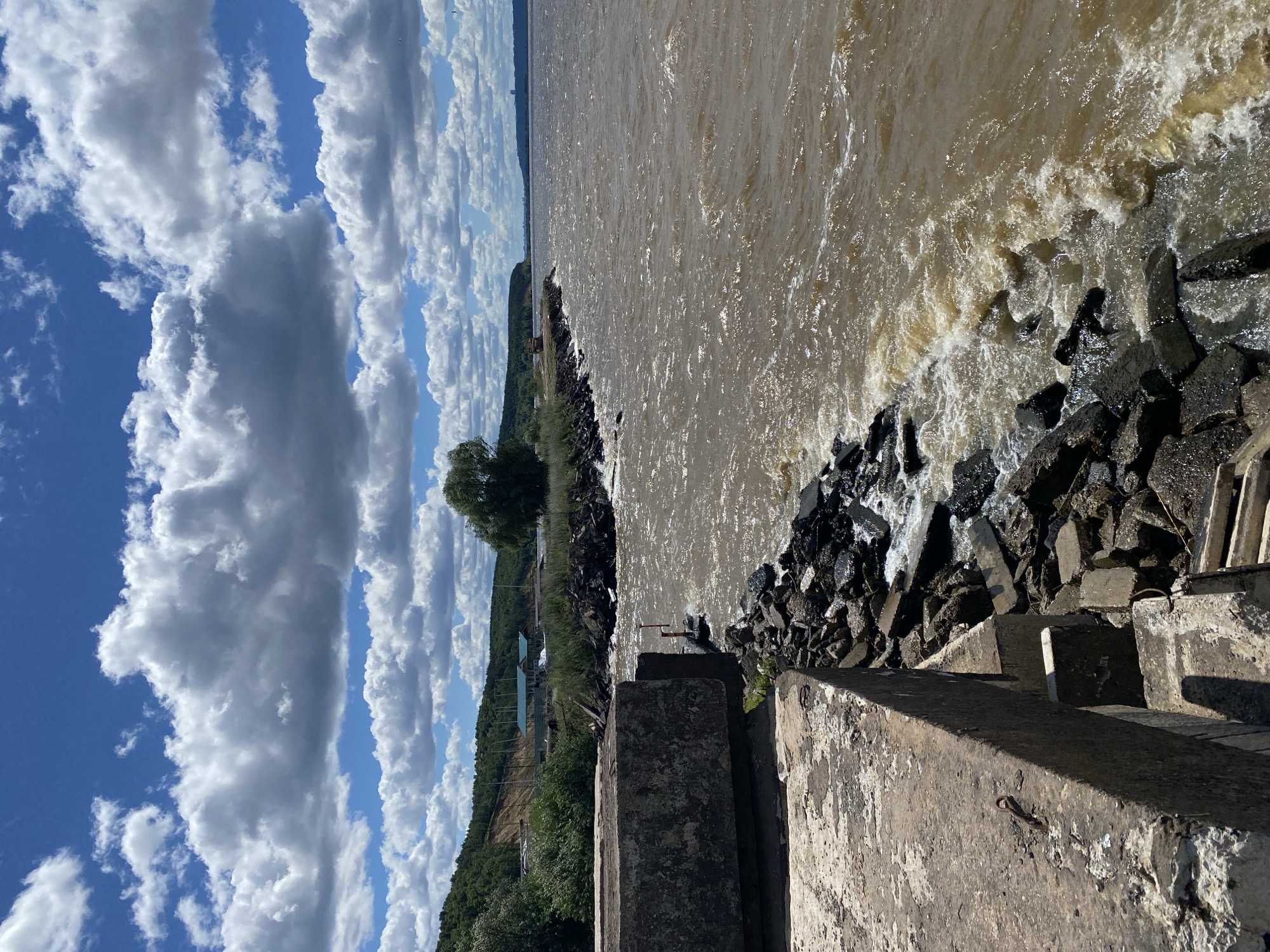 The image depicts a view of the Volga River in Leninsky, Russia, on a cloudy day. The foreground features a weathered concrete structure, possibly a pier or embankment, with visible rebar. Large rocks and rubble form a jagged edge along the water's interface with the concrete. The river itself is a muddy brown color, with choppy waves and white foam indicating some turbulence. A rocky shoreline extends into the distance, bordered by lush green vegetation and a gently sloping hill covered in trees. Along the distant shoreline, there are several buildings with light-colored roofs and a few masts, suggesting a small settlement or harbor. The sky is a mix of bright blue patches and large, white cumulus clouds, some of which cast shadows. The overall scene is natural and somewhat rugged, with a sense of expansive water and a clear, albeit partially overcast, sky.