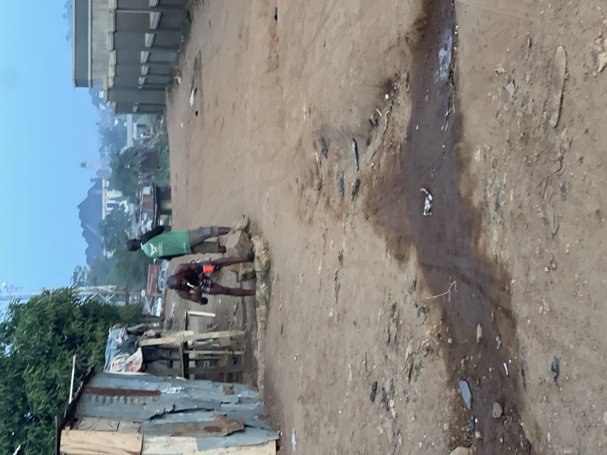 This image captures an outdoor scene in a developing urban or semi-urban area, consistent with parts of Abuja, Nigeria. The ground primarily consists of dry, reddish-brown dirt and sand, uneven and scattered with various pieces of litter, including plastic fragments and other debris. A visible trail of murky water or mud runs through the right side of the foreground and mid-ground, suggesting poor drainage or recent rainfall.
Two men are the main subjects in the mid-ground. The man on the left wears a bright green t-shirt and light-colored trousers, standing upright and looking forward. Beside him, to the right, another man is bare-chested, wearing patterned shorts, and appears to be looking downwards or engaged with an orange object held in his hands, possibly a cup or container.
On the far left of the frame, a makeshift structure constructed from corrugated metal sheets and wooden planks is visible, typical of an informal dwelling or small business. Behind this structure and further into the background, vibrant green trees and dense foliage add natural elements to the landscape. In the distant background, under a pale blue, hazy sky, several multi-story buildings can be seen. Some appear to be residential, while others might be under construction or development, indicating an evolving urban environment. The soft, diffused light suggests either early morning or late afternoon. There is no visible text in the image.