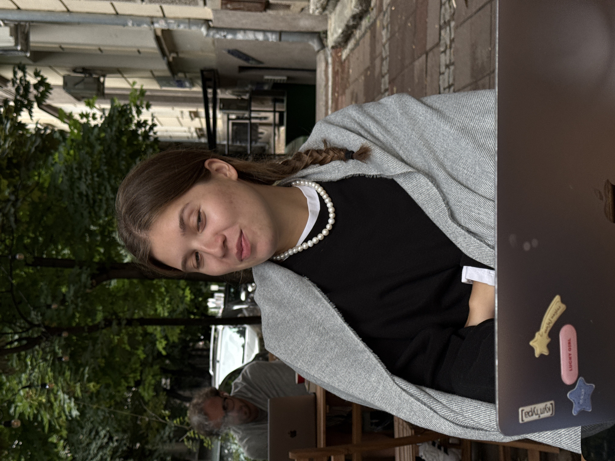 A young woman with a braided ponytail sits at an outdoor cafe in Belgrade, Serbia, wrapped in a gray blanket and wearing a pearl necklace. She is looking to her left with a slight smile. In front of her is a laptop adorned with stickers, including one that reads "LUCKY GIRL". In the background, a man with glasses is visible, also working on a laptop at another table. The cafe appears to be situated on a street with trees and buildings lining the right side of the frame.