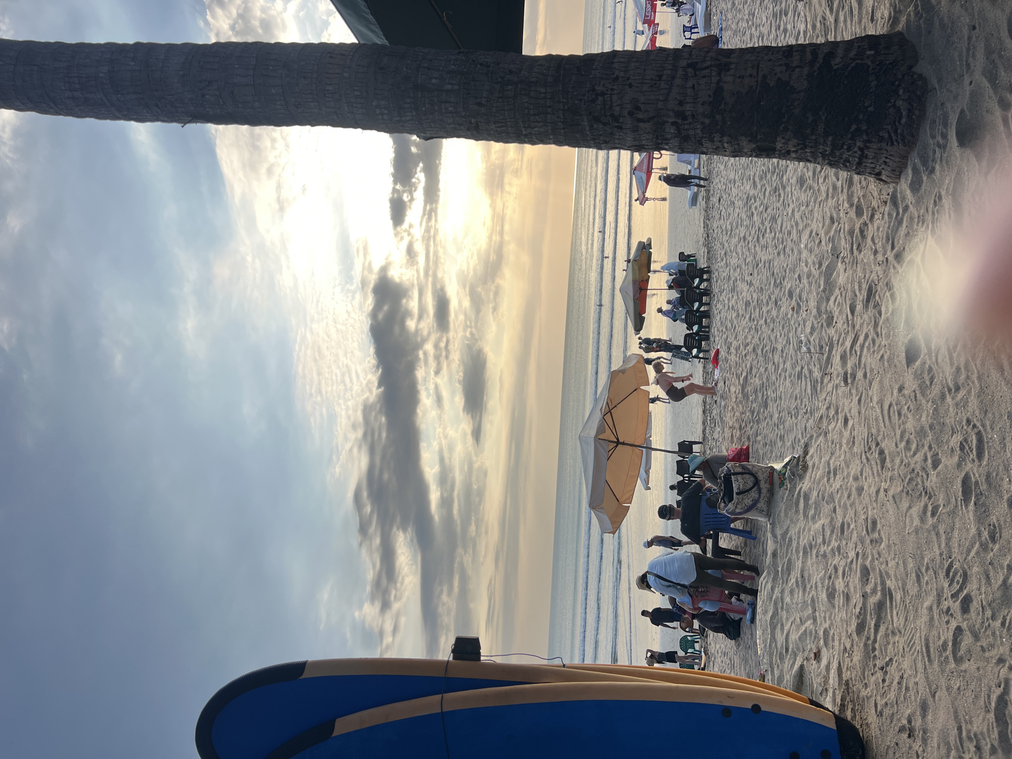 The image depicts a serene beach scene at sunset in Kuta, Indonesia. In the foreground, a large surfboard stands vertically on the left, its blue and yellow surface reflecting the golden light. The sandy beach is populated with several individuals and groups. Some are seated under umbrellas, enjoying the view, while others are standing or walking. A tall palm tree trunk dominates the right side of the frame, its rough texture a stark contrast to the smooth sand. In the middle ground, the ocean stretches out to the horizon, with gentle waves lapping at the shore. The sky is filled with a dramatic display of clouds, illuminated by the setting sun, casting a warm, ethereal glow over the entire scene. The overall atmosphere is one of relaxation and natural beauty.