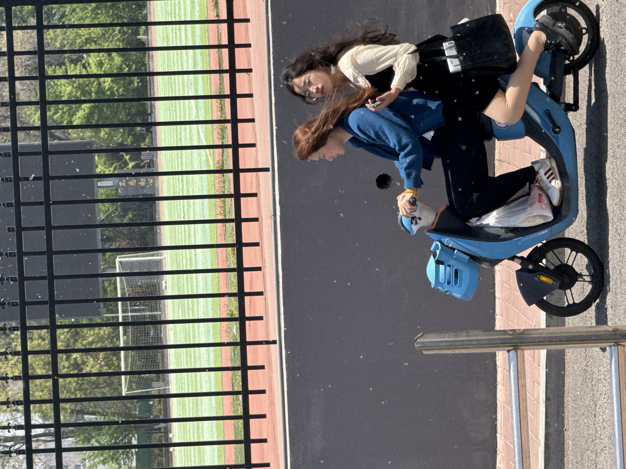 Two young women are on a light blue electric scooter, which is parked on a gray paved surface next to a fenced sports field. One woman with dark hair and glasses sits behind the other, looking at a phone. The front woman has reddish-brown hair and wears a blue sweater, and is facing towards the sports field. The field has a soccer goal at one end and a running track border. The location is presumed to be Zhongtun, China.