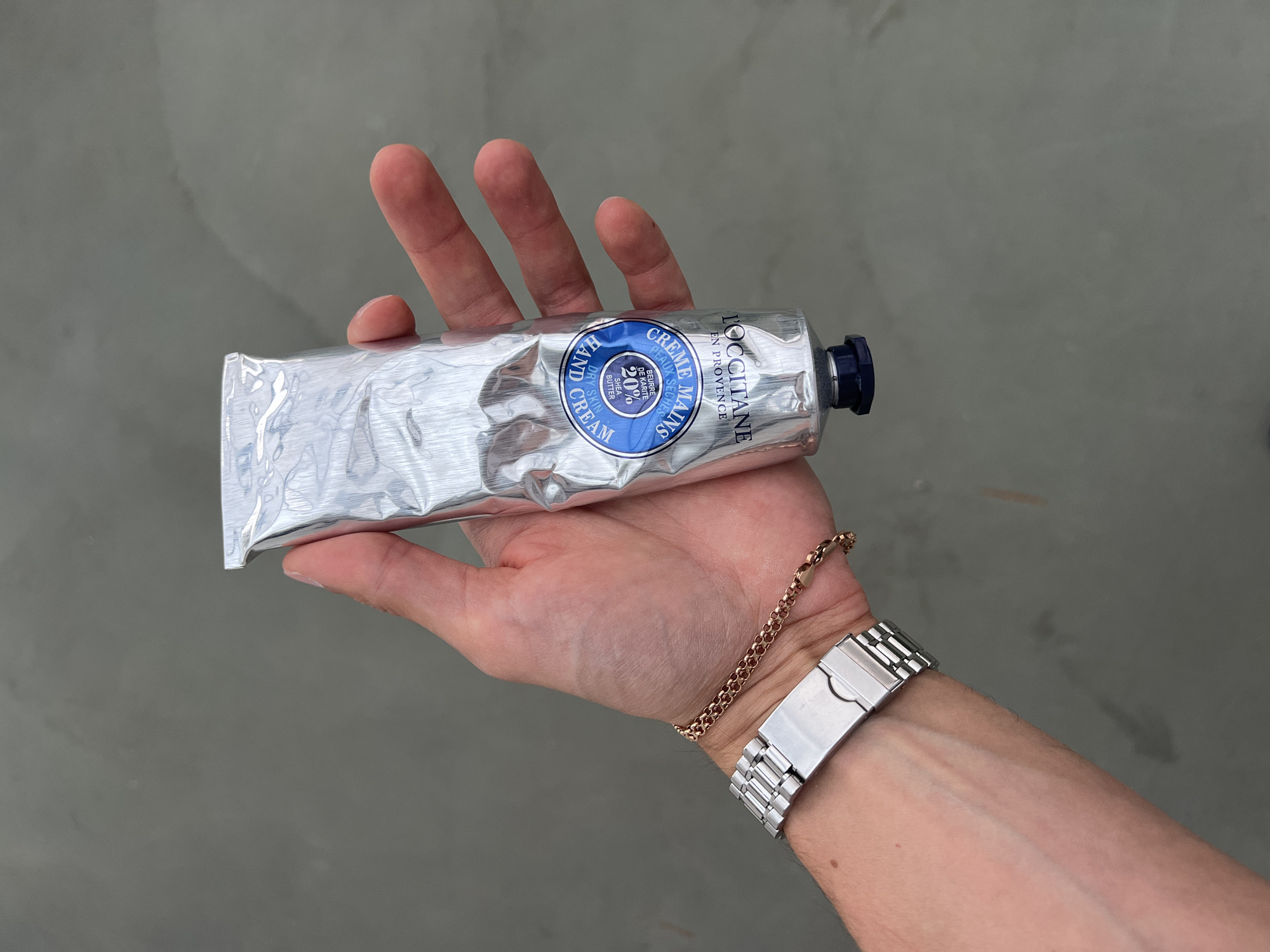 An individual's hand is holding a silver tube of L'Occitane Shea Butter Hand Cream. The hand is adorned with a silver watch and a thin rose gold bracelet. The background is a plain, gray, textured surface, possibly concrete, with subtle lighting variations. The image does not provide any information about the city of Borowa, Poland.