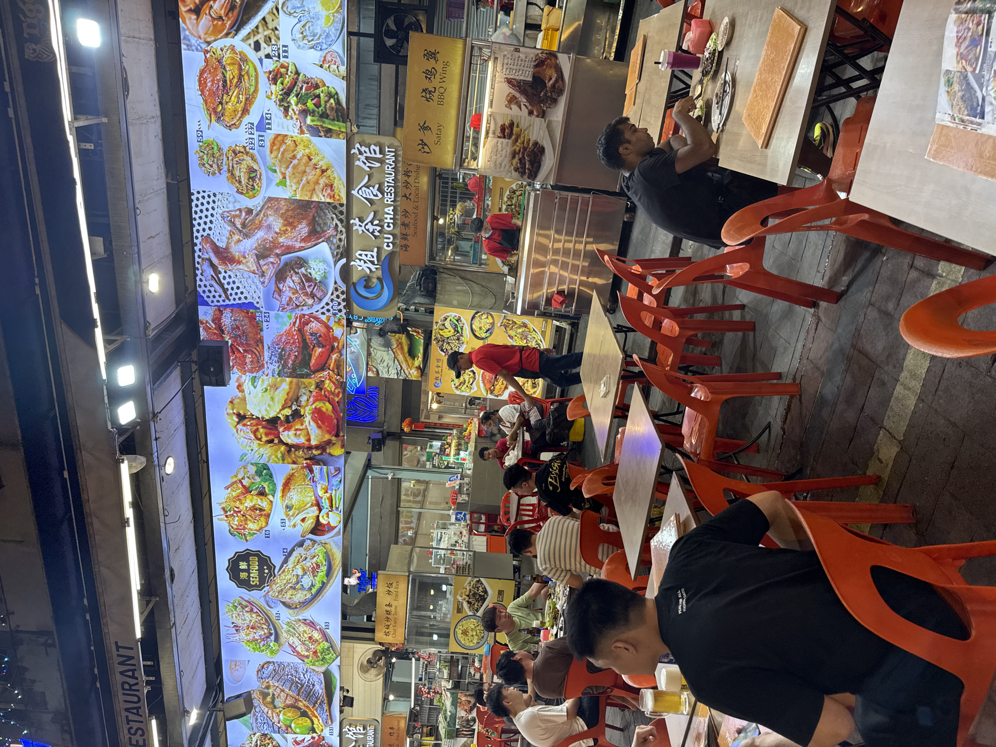 This image captures a vibrant outdoor night market or hawker center, likely in Kuala Lumpur, Malaysia, bustling with diners and food stalls. The scene is illuminated by bright artificial lights, suggesting nighttime. Numerous people, primarily men, are seated at simple square tables with distinctive orange plastic chairs, engaged in eating, drinking, and socializing. Many are casually dressed. In the foreground, a man in a black t-shirt is seen from behind, holding a glass of what appears to be beer.
Dominating the left side of the frame are large, brightly lit vertical menu boards displaying a wide variety of seafood and local dishes with photographs and prices. Visible text on these boards includes "ESTAURANT," "海鲜 SEAFOOD," "炸串 Fried Skew," and "槟城炒粿条炒饭 Char Kuey Teow Fried Rice." Several horizontal signs further identify specific food stalls, such as "CU CHA RESTAURANT" (粗茶食馆), which advertises "Seafood & Local Dishe," alongside offerings like "沙爹 Satay" and "烧鸡翼 BBQ Wing."
Food preparation counters and display cabinets are visible behind the dining area, with staff members in red or black t-shirts attending to customers. The atmosphere appears relaxed and lively, typical of a bustling street food environment. Scattered on the tables are plates, utensils, and beverage glasses, including one with a "Tiger" beer logo. The combination of diverse local cuisine, Chinese characters, and the general setup strongly points to a Southeast Asian dining experience.