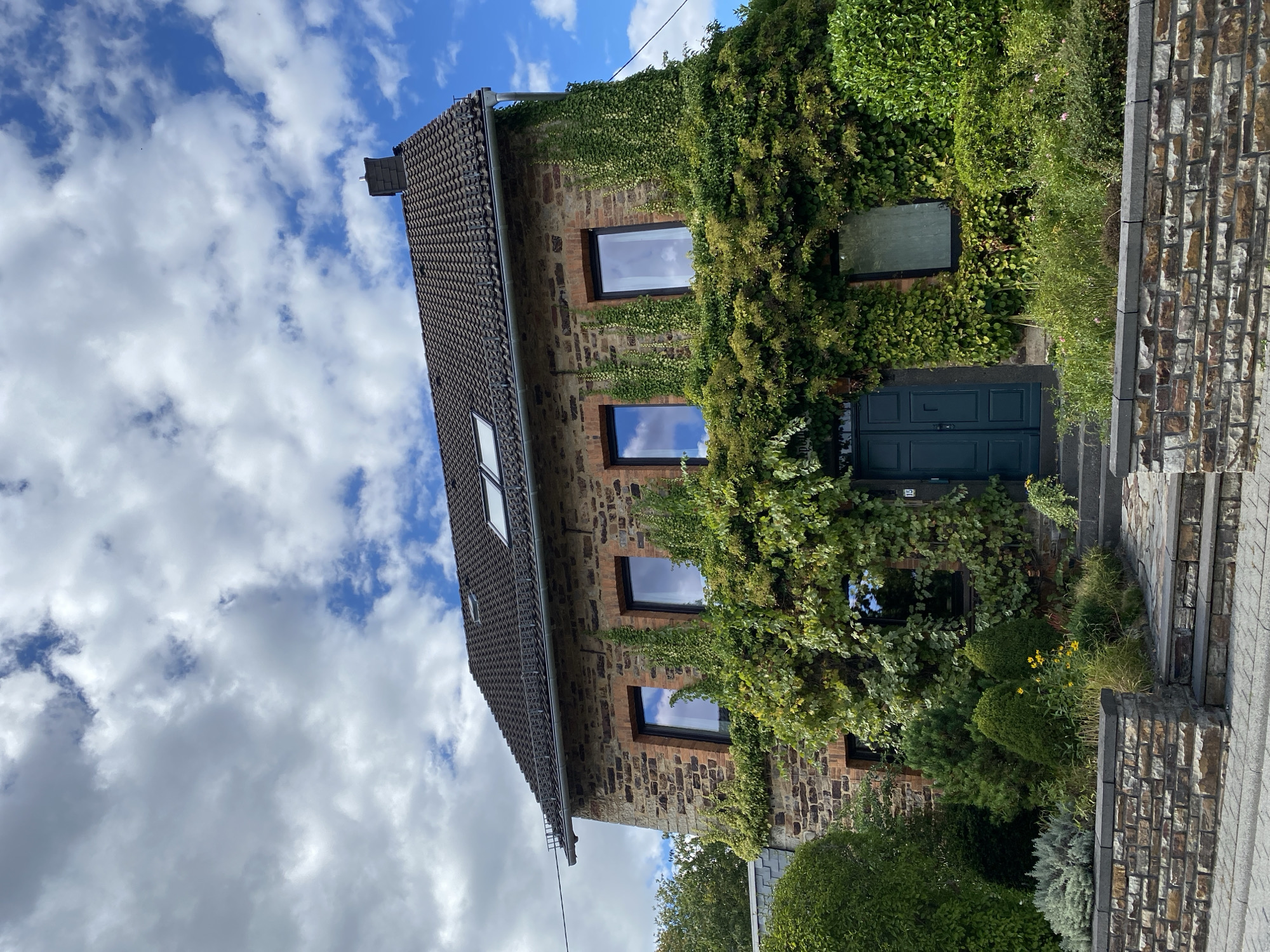 The image shows a two-story stone house covered in ivy, located in Flammersfeld, Germany. The house has a dark tiled roof with two skylights and a chimney. It features several windows with dark frames on both floors. The front entrance is a dark teal double door, accessible by a set of stone steps. The area in front of the house is landscaped with various green shrubs, small trees, and yellow flowers. A low stone wall borders the property along the street, which is paved with cobblestones. The sky is partially cloudy with patches of blue. There are no people visible in the image.