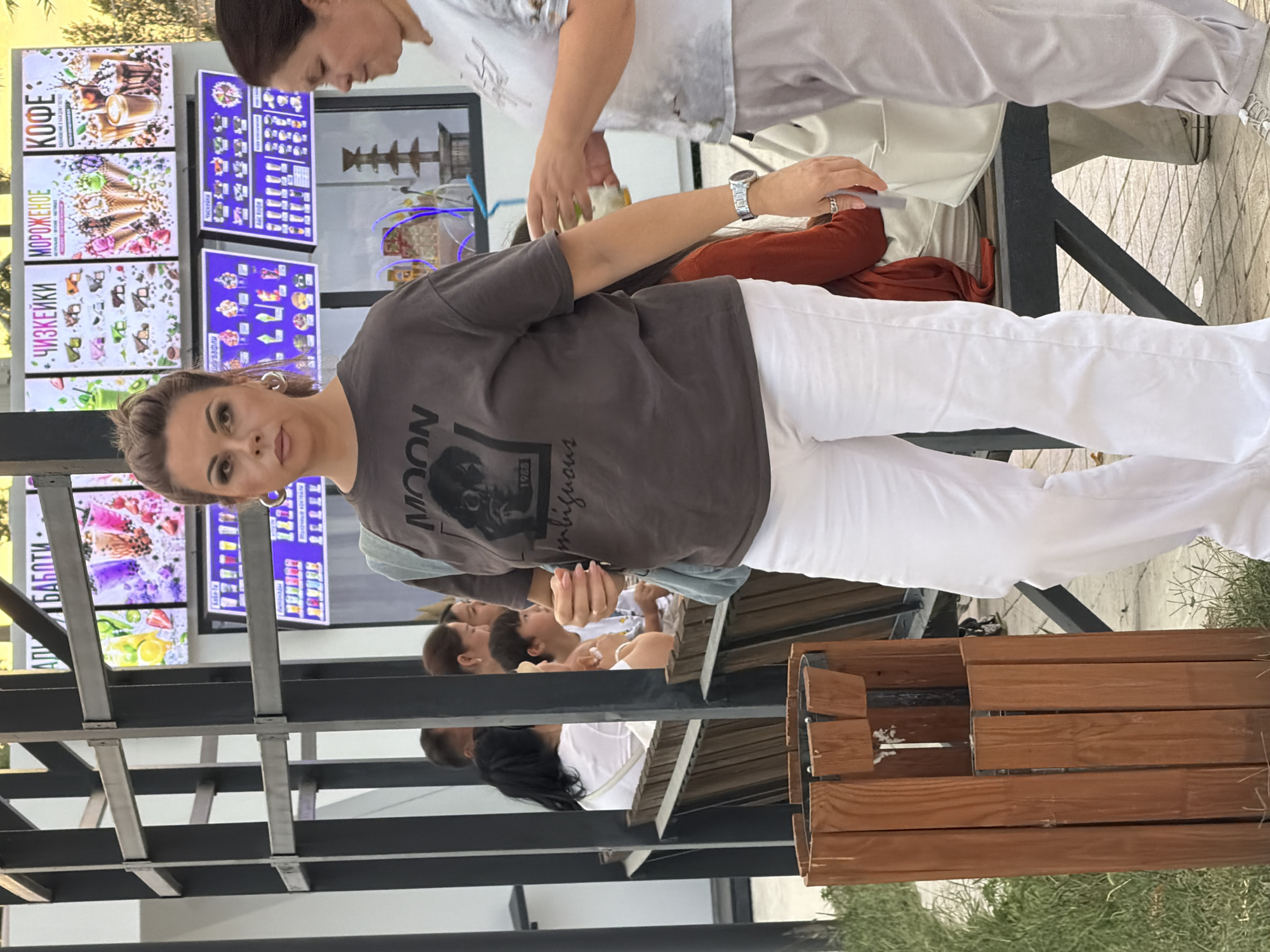 In a city setting, a woman with dark hair pulled back stands in the foreground, facing the camera. She is wearing a dark gray t-shirt with a graphic print and white pants. A light blue strap of a bag is visible over her shoulder. To her right, another woman in a light-colored shirt is partially visible, appearing to hand something to the woman in the foreground, though their interaction is unclear. Behind them, a menu board displays various food and drink options, including ice cream and coffee, with Cyrillic text. Several other people are seated on a wooden bench in the background, suggesting a public area like a cafe or food stall. A wooden trash receptacle is positioned to the left of the main subject. The environment appears to be outdoors, with a paved ground and greenery visible.