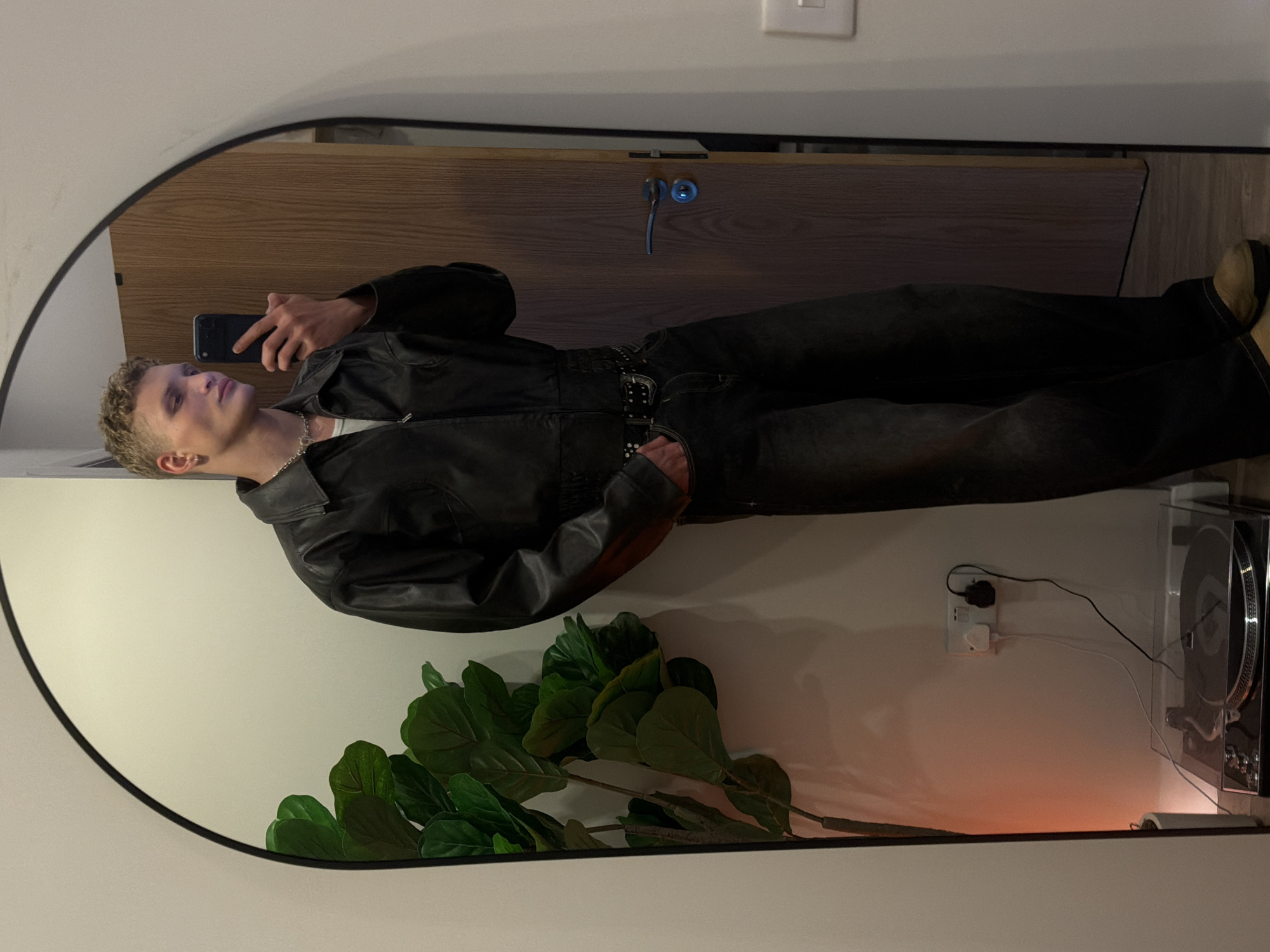 An indoor mirror selfie captures a young man standing in what appears to be a room in London, United Kingdom. He has short, curly, light-colored hair and is wearing a dark leather jacket over a white t-shirt, dark trousers, and a chain necklace. He is holding a dark smartphone, likely an iPhone, to take the picture, with a calm, composed expression.
The full-length, arched mirror reflects a scene that includes a wooden door with a silver handle and lock mechanism behind the man. To the right, a large artificial plant with dark green leaves sits on the floor. Further right, a white UK-style wall outlet with multiple plugs and wires is mounted on a light-colored wall. Adjacent to the outlet, a silver and black record player with a clear dust cover is visible. A warm, orange-hued light source near the floor casts a soft glow across the room. The man's right foot, wearing a light brown boot or shoe, is visible at the bottom of the frame, and he has a studded dark belt. The lighting suggests an indoor setting, possibly during the evening. No discernible text is visible.