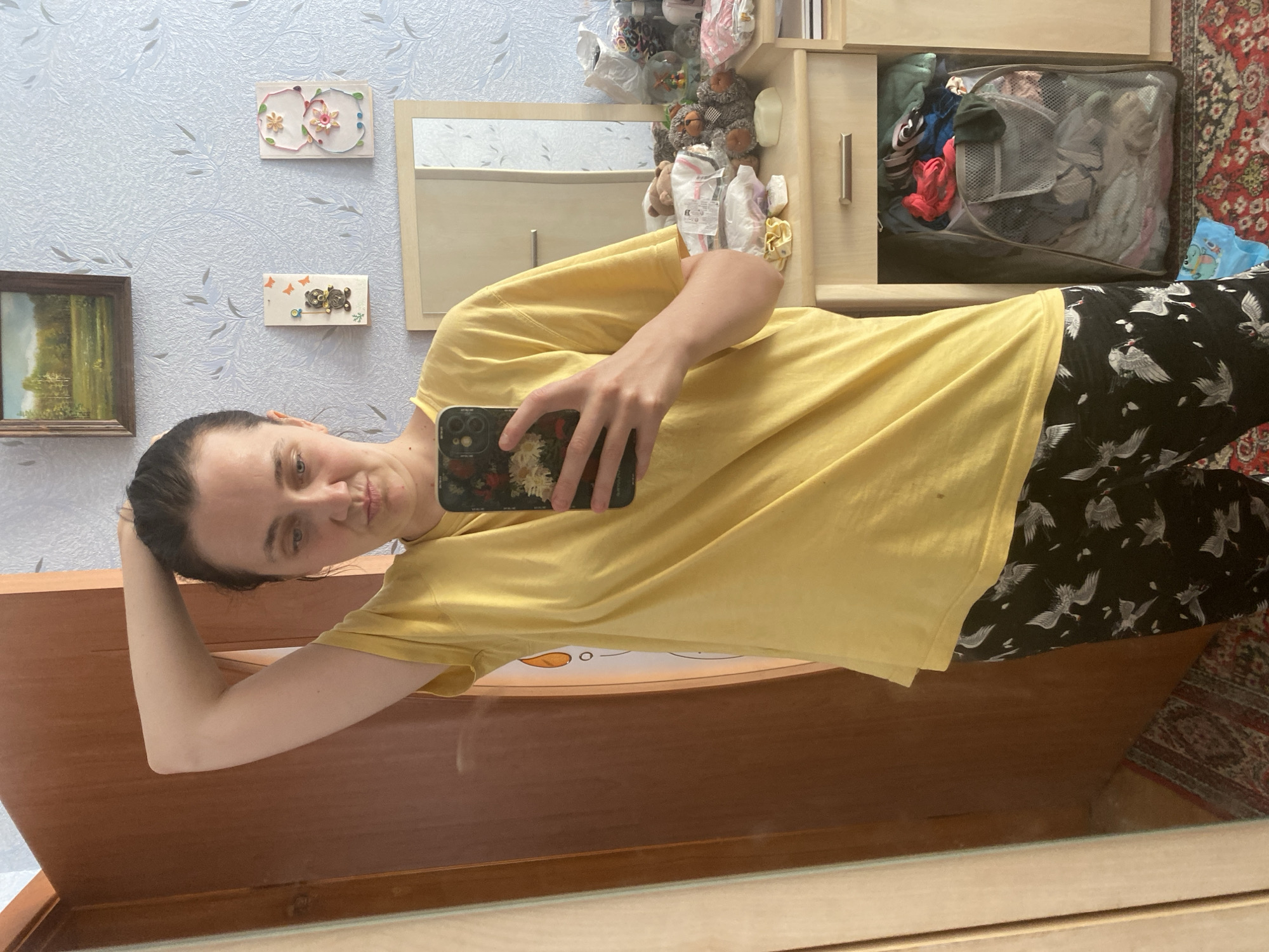 A young woman, with dark hair pulled back and her arm raised, stands in front of a mirror, taking a selfie with her phone. She is wearing a loose, bright yellow t-shirt and black pajama pants with a white crane pattern. Her expression is somewhat pouty. The background reveals a room with patterned wallpaper, a small framed landscape painting, and decorative items on the wall. To the right, a wooden dresser with a mirror is visible, topped with various items including stuffed animals and toiletries. Below the dresser, a laundry hamper filled with clothes is positioned on a patterned rug. The scene appears to be an indoor setting, possibly a bedroom.