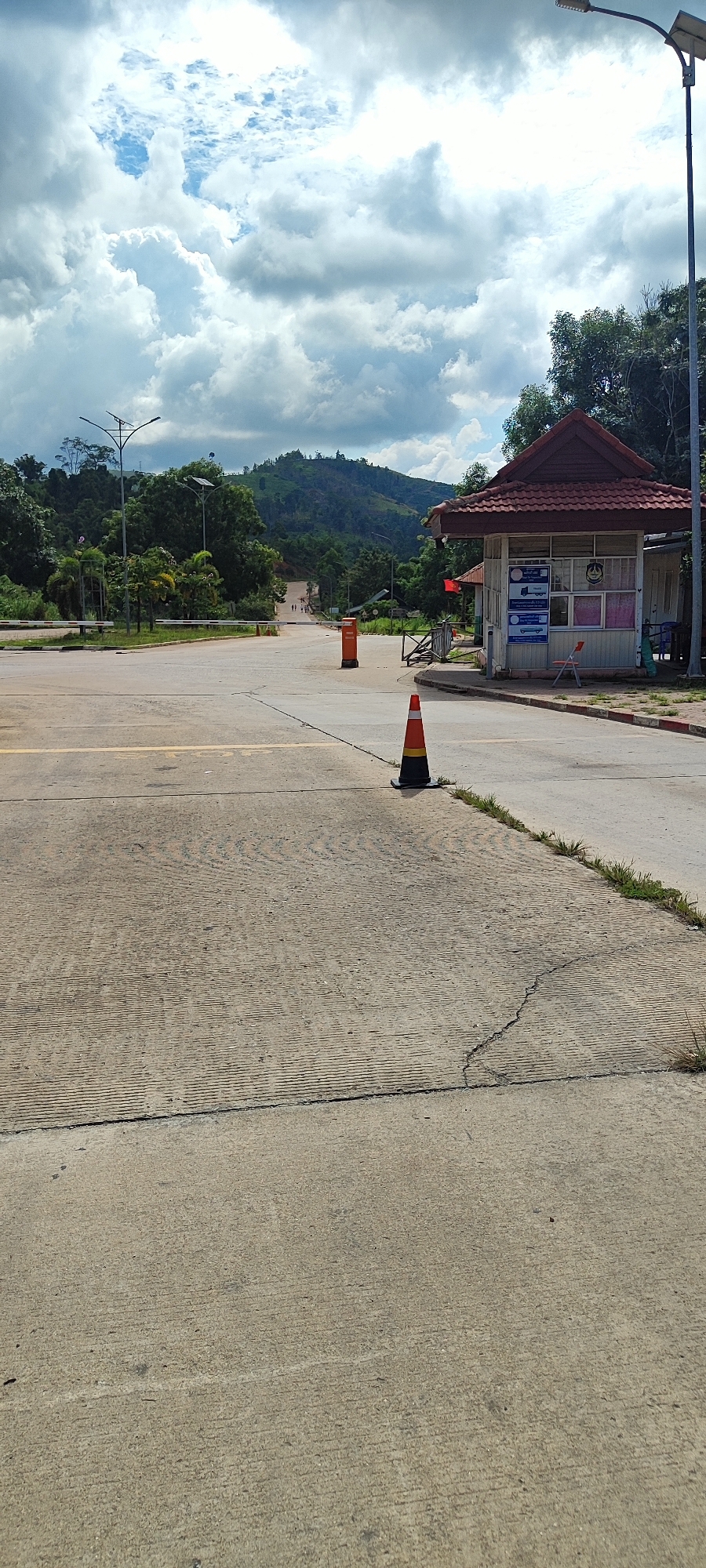 This is a vertical, eye-level, full shot of a border crossing in Xayden, Laos. The scene is dominated by a cloudy sky and a winding road leading into a hilly, tree-covered landscape. In the foreground, a concrete road is visible with yellow lines and a "STOP" marking. Two orange traffic cones are placed on the road, one near a small security building on the right, and another further down the road. The security building, with a red-tiled roof, appears to be staffed, with a flag and signs visible. A barrier gate is also present in front of the building. In the distance, several people are walking along the road, and vehicles are parked. The overall impression is one of a moderately busy border crossing with a backdrop of lush green hills under a dramatic sky.