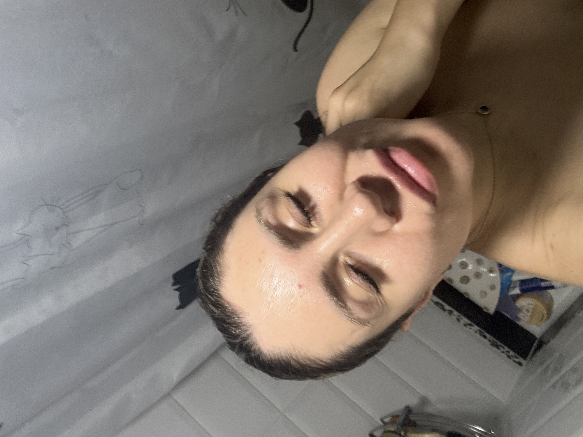 A woman is taking a selfie in a shower, her face is close to the camera and her eyes are closed. She is wearing a necklace. The background shows a shower curtain with cat drawings and white tiled walls. The scene is indoors and appears to be in a residential bathroom.