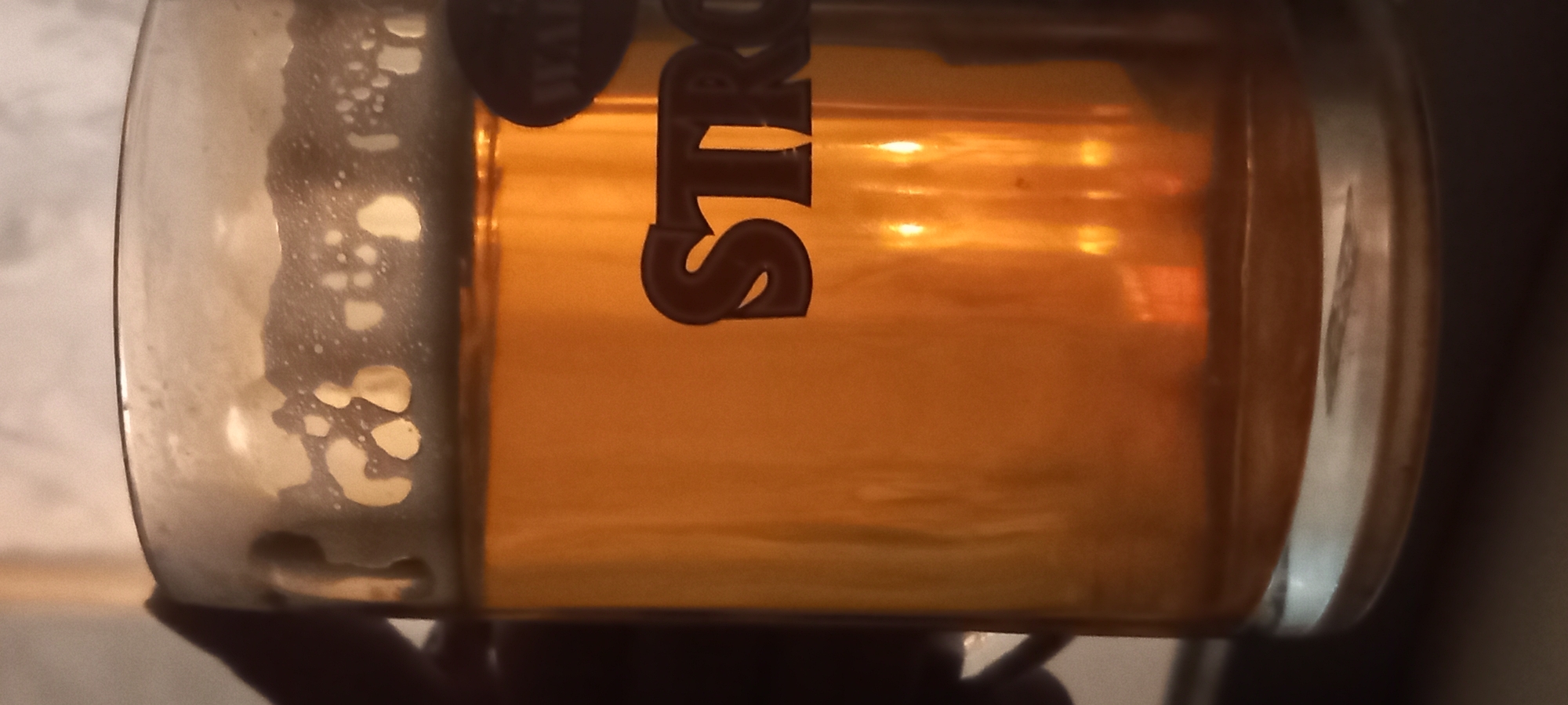 A close-up shot shows a glass mug filled with golden-colored liquid, likely beer, with a frothy head. The mug has the word "STRONG" embossed on it in a dark, stylized font. A dark shadow, presumably a hand, is visible holding the mug from the left. The background is out of focus, with a light-colored, textured surface and some reflections of warm light. There is no visible indication of the city of Kryvyi Rih, Ukraine.