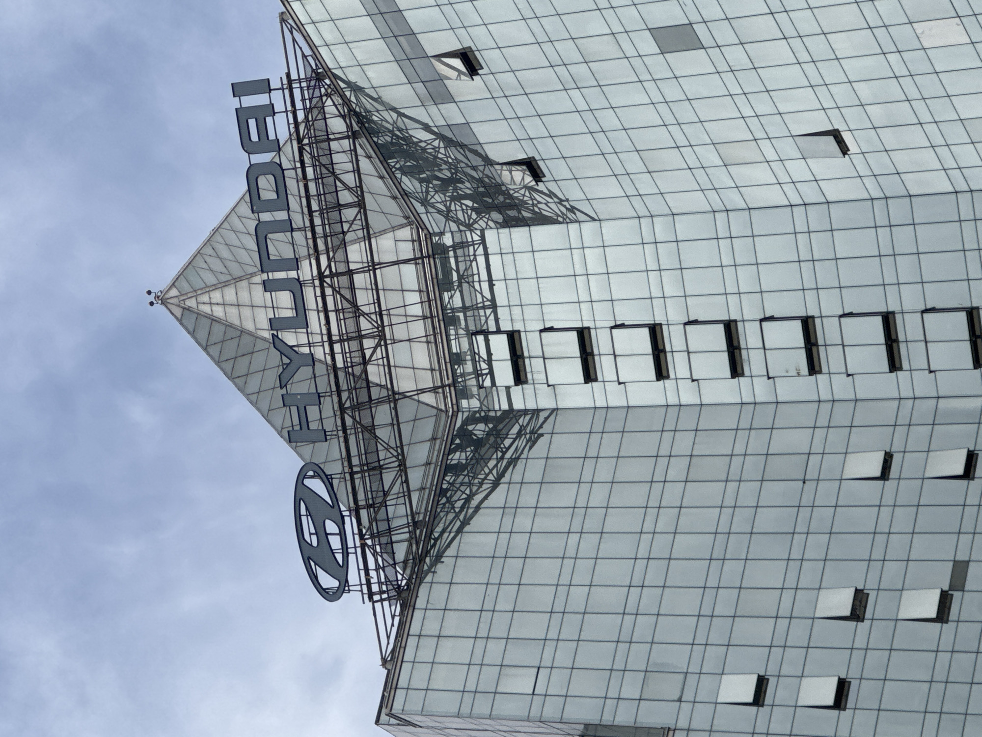 A low-angle, exterior medium shot shows the upper portion of a modern, glass-facade building against a pale, overcast sky. The building features a prominent, pyramid-shaped glass structure at its apex, topped by the dark gray, metallic Hyundai logo and lettering. The building's facade is composed of numerous rectangular glass panes, reflecting the sky and creating a grid-like pattern. Several windows on the building are open, tilted upwards. The overall impression is one of architectural grandeur and corporate presence in an urban setting, specifically identified as Almaty, Kazakhstan.