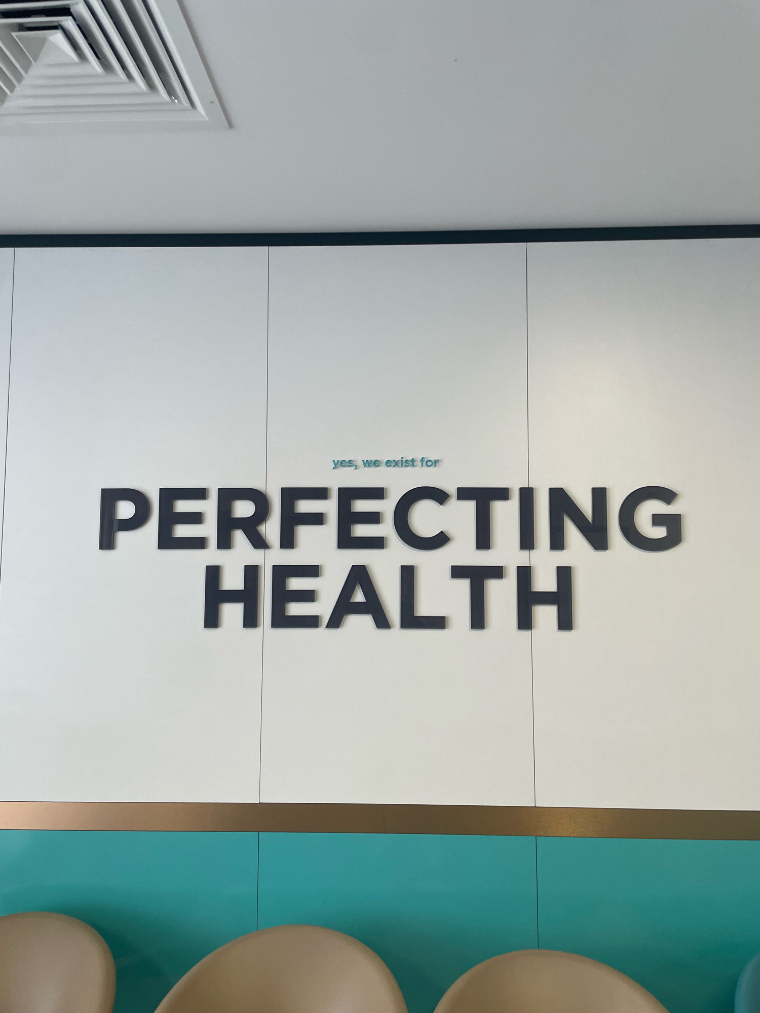 The image shows a wall with the words "PERFECTING HEALTH" written in large, black, three-dimensional letters. Above these words, in smaller, light blue letters, it says "yes, we exist for". The wall itself is white and appears to be divided into panels. Below the white wall is a horizontal brass-colored strip, and beneath that, a teal-colored wall. The bottom of the image shows the tops of several light brown chairs. In the upper left corner, a white air vent is visible against a white ceiling. The setting appears to be an indoor waiting area, possibly a clinic or wellness center. No people are visible in the frame.