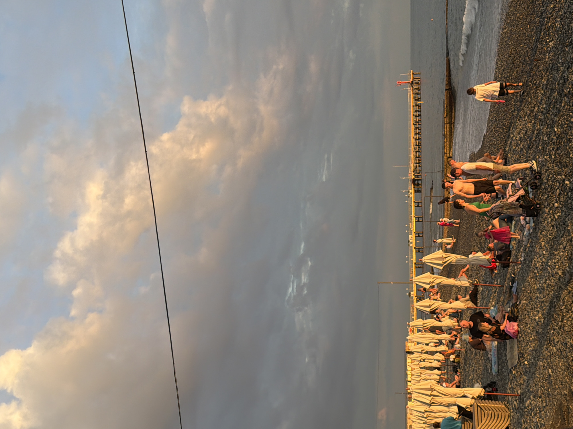 The image shows a crowded pebble beach in Sochi, Russia, during what appears to be sunset. The sky is filled with dramatic, colorful clouds, ranging from light yellow and orange to dark gray. A thin black wire stretches across the upper part of the frame, possibly a power line or telephone wire. In the background, a long pier with yellow railings extends into the calm sea, topped with a red and white navigation beacon. Along the shore, numerous white beach umbrellas are set up, some open and some closed, with people scattered among them. The beach is populated with individuals and small groups of people, some sitting on towels, others standing or walking. The overall atmosphere suggests a leisurely evening by the sea.