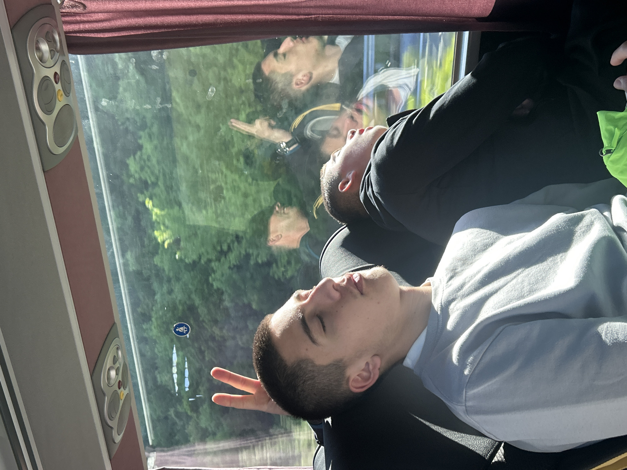 A young man sleeps against a seat on a bus or train. His head leans against the headrest. He wears a light-colored sweatshirt. Another man is sleeping behind him, wearing a dark jacket. The window shows a reflection of people outside, as well as a view of trees and vegetation. The interior of the bus or train is visible, with window trim and controls. There is a blue handicap symbol on the window. Without additional information, it is impossible to confirm that the image was taken in the Municipal Unit of Kato Olympos, Greece.