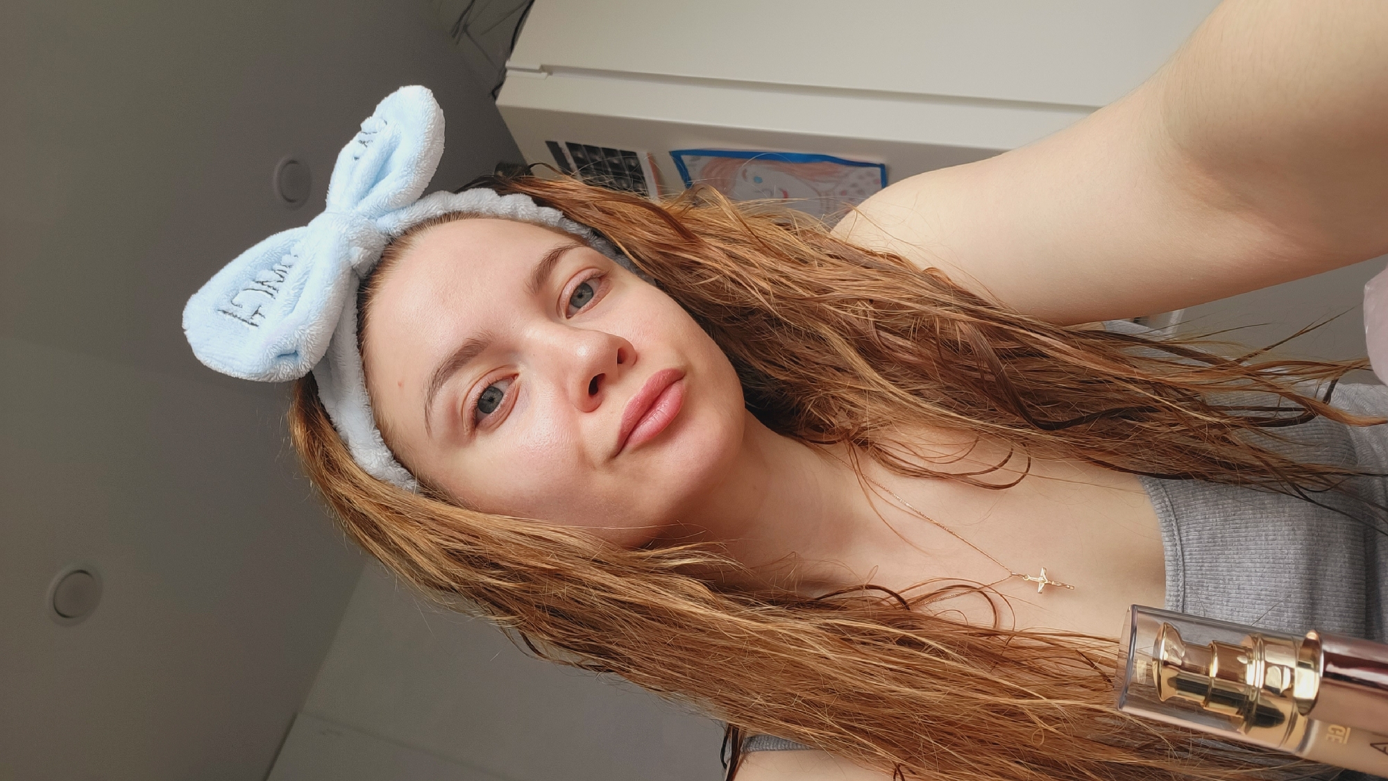 A young woman with light skin, light eyes, and wet reddish-brown hair wears a light blue headband with bunny ears. She is positioned at an angle, taking a selfie with her right arm extended. She is wearing a gray tank top and a delicate gold necklace with a small pendant. A bottle of cosmetic product, with a transparent container and gold accents, is visible in her hand. The background includes a white wall with a light switch and what appears to be a child's drawing. The scene is indoors, likely in a bathroom or bedroom. There is no specific information to confirm it is in the city of Chaikovsky, Russia.