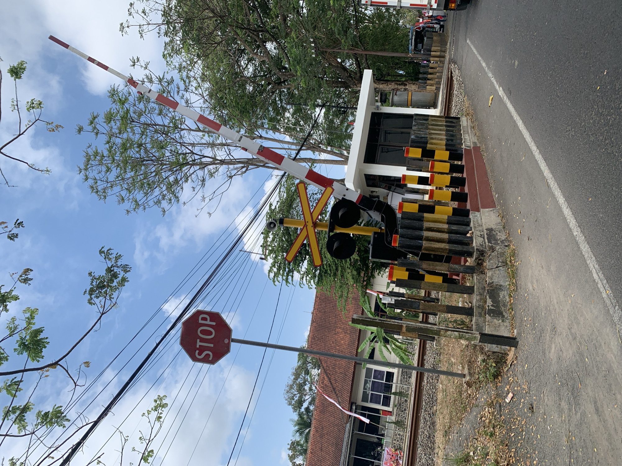 This is a photograph taken at a railway crossing in Ciamis, Indonesia. The image features a red octagonal stop sign positioned on a metal pole, with the word "STOP" clearly visible in white letters. A railway crossing barrier, painted with alternating red and white stripes, is raised, indicating that trains are not currently passing. The barrier is positioned next to a small white control booth. The railway tracks are visible in the foreground, alongside a paved road. Several poles with black and yellow stripes mark the boundary of the crossing. In the background, a building with a terracotta tiled roof is visible, along with lush green trees and a bright blue sky with scattered clouds. The overall scene depicts a functional railway crossing in a moderately vegetated urban or semi-urban environment.