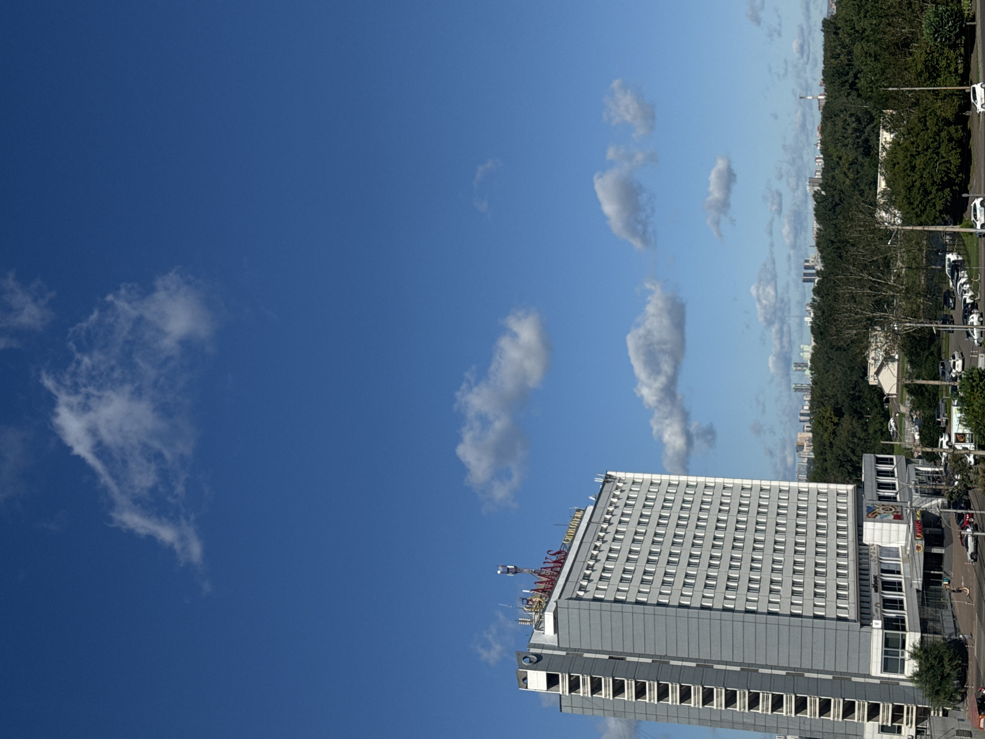 The image depicts a view of the "AMAKS Hotel" in Krasnoyarsk, Russia. The hotel is a tall, grey, multi-story building with numerous windows and balconies. It is situated near a dense green forest and appears to be in an urban or semi-urban environment, with other buildings and a road visible in the foreground. The sky above is bright blue with scattered white clouds. There are cars parked in the lot in front of the hotel. The hotel has signage that reads "AMAKS" in red lettering at the top and other text in Cyrillic script on its lower levels. The overall scene suggests a sunny day in Krasnoyarsk.