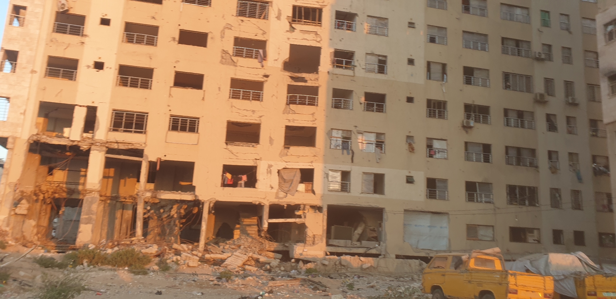 The image shows a partially destroyed apartment building in Gaza City, Palestinian Territories, bathed in the warm light of what appears to be sunset or sunrise. The building exhibits significant structural damage, with multiple floors showing collapsed sections, exposed rebar, and shattered windows. Debris, including concrete rubble and twisted metal, litters the ground in front of the building and the surrounding area.
Several windows on the intact sections of the building are open or broken, and some have clothes hanging from them, suggesting that the building may have been inhabited or recently abandoned. Balconies with metal railings are visible on various floors, some appearing intact and others damaged. Air conditioning units are mounted on some exterior walls.
In the foreground, two yellow pickup trucks are parked. The truck on the left is a small, older model with its tailgate down. The truck on the right is partially obscured by tarps. The ground is uneven and covered with rubble, sand, and some sparse vegetation. The overall scene conveys a sense of destruction and hardship.