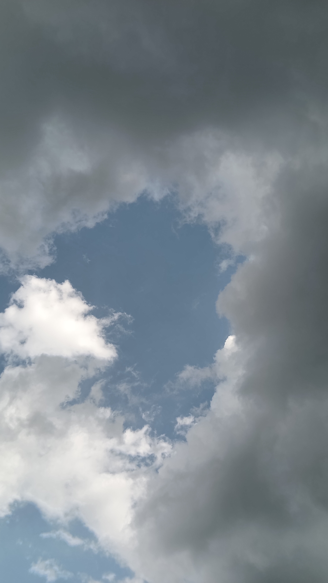 The image shows a patch of light blue sky visible through a break in the cloud cover. The clouds surrounding the opening vary in color from white to dark gray. The location cannot be determined from the image.
