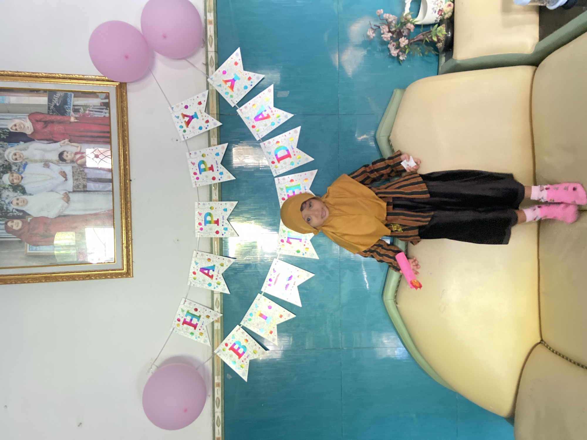 A girl wearing a hijab and pink socks is lying on a light yellow couch, holding a pink toy. The room is decorated for a birthday, with pink balloons and a "HAPPY BIRTHDAY" banner on the wall. A framed picture hangs on the wall. The scene is in Gelam, Indonesia.