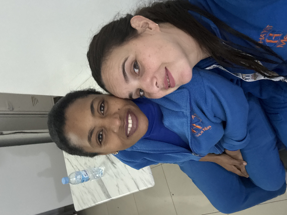 This image, taken in Meknes, Morocco, features two women posing together indoors, showcasing a warm and friendly interaction. The photo is oriented as if the subjects are leaning towards the right.
The woman positioned in the foreground on the left has a dark complexion, dark hair neatly pulled back, and a radiant, wide smile. She is dressed in a blue top, likely part of a tracksuit or comfortable uniform, which has an orange embroidered logo on her left chest, appearing to read "Soumia" or "Somia" with a stylized design.
Behind her, on the right, is another woman with a lighter skin tone, long dark hair, and light freckles. She offers a gentle smile and rests her head affectionately on the shoulder of the first woman. Her blue top matches the other's and also features orange embroidered text on her right chest, which is partially visible and appears to spell "HAYT ME".
Both women's blue attire, seemingly made of a fleece-like material, suggests they might be wearing uniforms, team wear, or matching casual outfits. Their expressions clearly convey happiness, affection, and camaraderie.
The background details include a light-colored table with a subtle marble pattern, located to the left of the women. On this table, a plastic bottle of "Sidi Ali" mineral water is visible. The setting appears to be an indoor space with plain light walls and a light-tiled floor, indicative of a common room, office, or classroom, illuminated by bright, likely daytime, lighting.
