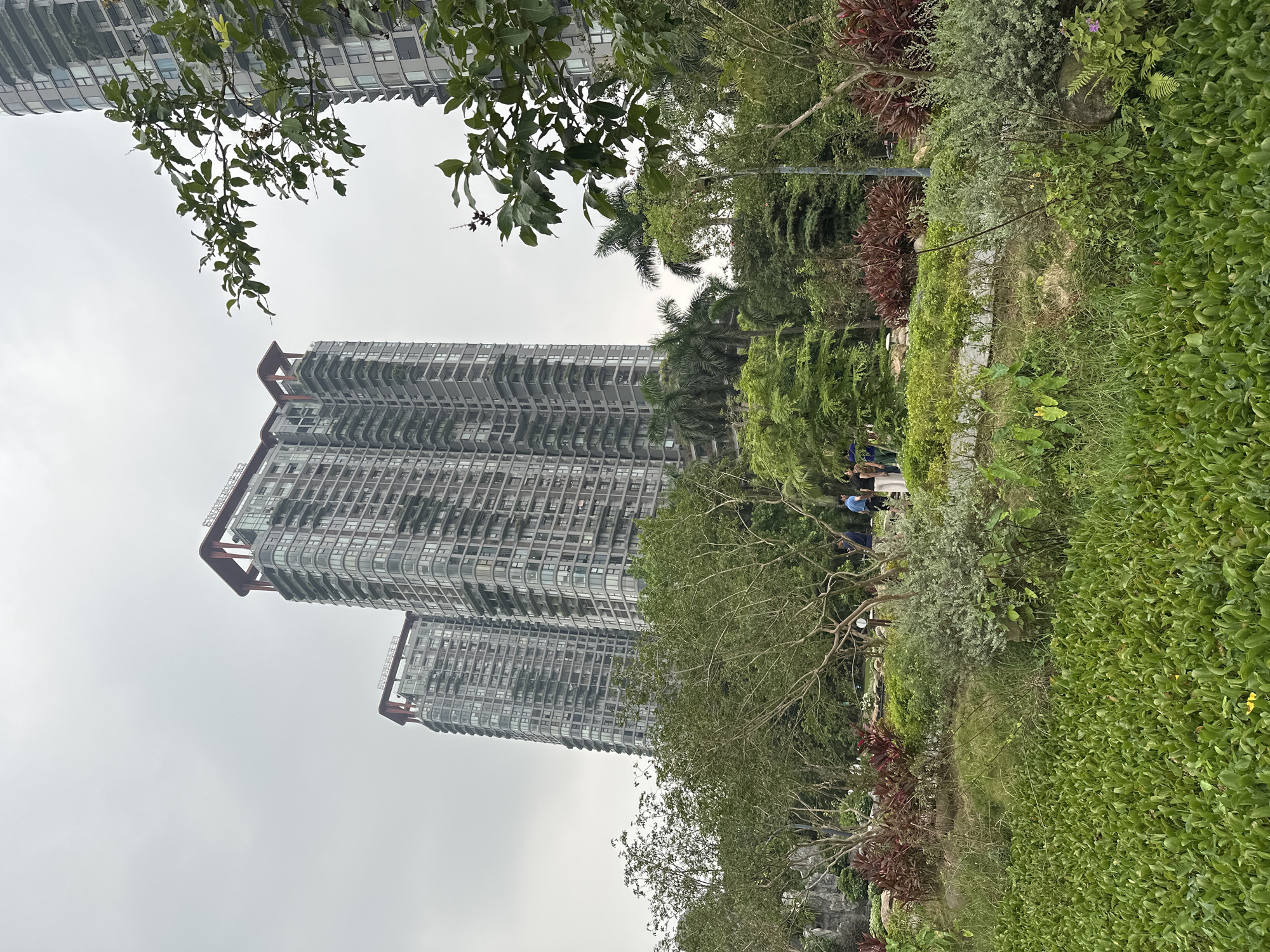 In the city of Phụng Công Commune, Vietnam, two modern, high-rise apartment buildings dominate the background against an overcast sky. The buildings are constructed with numerous windows and feature distinctive red architectural elements at their rooftops. In the foreground, a lush green park area is visible, characterized by dense foliage, trees, and a water body covered in aquatic plants. A paved pathway winds through the park, and several individuals are seen walking and interacting within the park space. Red and green ornamental plants are also present in the park, adding color to the landscape.