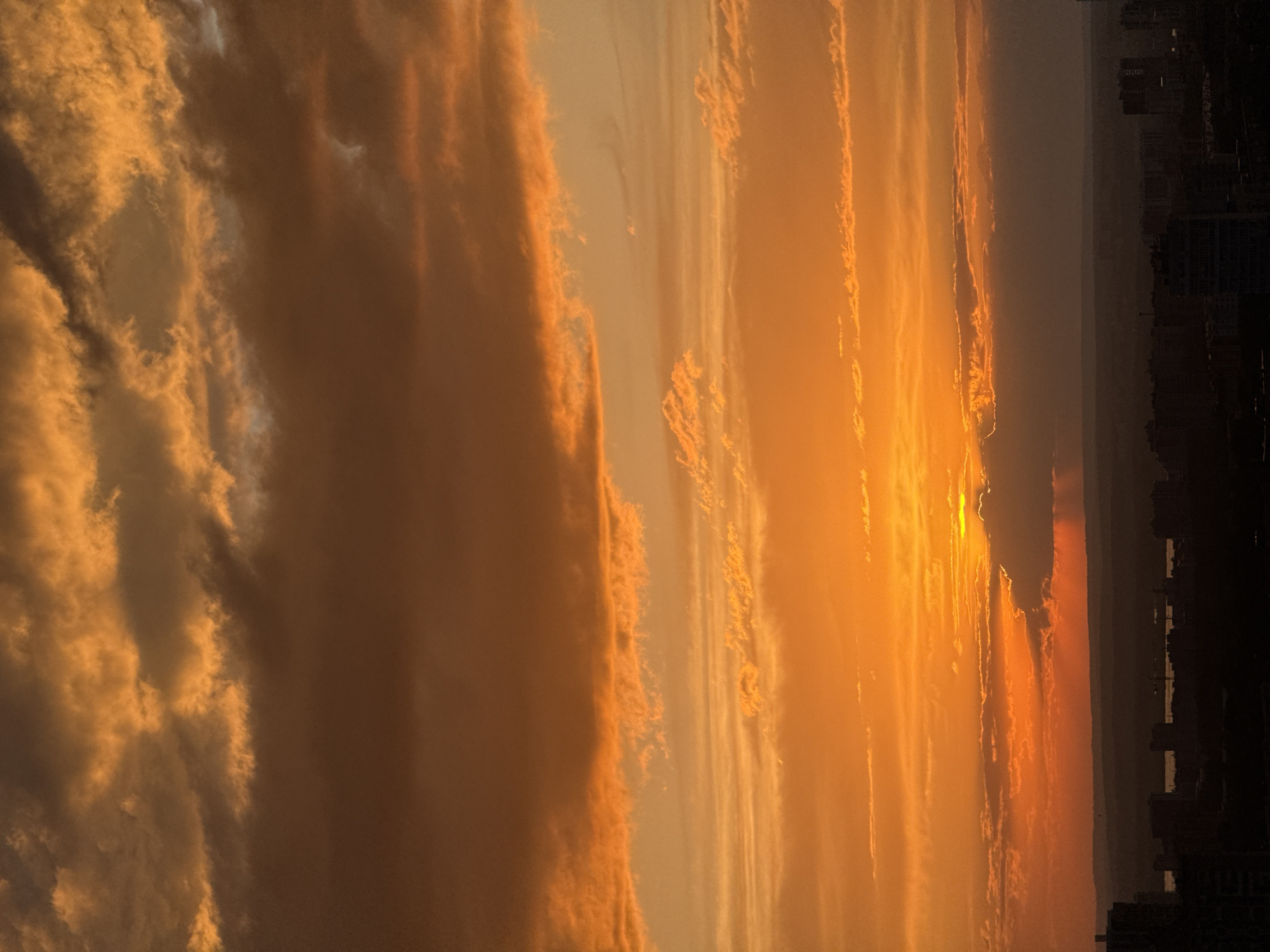 The image depicts a sunset over the city of Perm, Russia. The sky is filled with golden and orange clouds, and the sun is visible near the horizon. The city skyline is visible in the distance, with buildings silhouetted against the sky. The photo is oriented vertically.