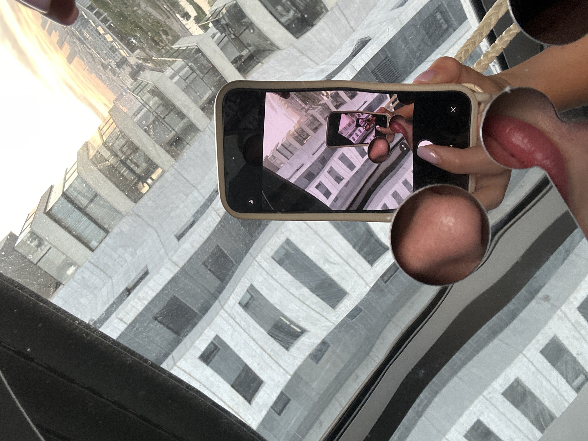 The image shows a hand holding a smartphone, which is taking a picture of a reflection. The reflection is of a modern building with many windows, seen from a high vantage point. The sky in the reflection appears bright, suggesting it's daytime.
The main subject is a person's hand, with manicured nails, holding a light-colored smartphone. The phone's screen displays a reflection of the surrounding environment, which is a city with tall buildings. In the foreground, part of another person's face is visible, specifically their chin and part of their mouth. The overall scene is a bit abstract due to the reflections and the unusual framing, with parts of the image appearing warped or distorted.
The setting appears to be indoors, possibly in a room with large windows overlooking a cityscape. The time of day is likely late afternoon or early evening, given the warm, golden light in the reflection of the sky. There are no specific weather cues other than the bright sky. The presence of the smartphone and the act of taking a photo indicates someone is documenting the view or the surreal reflections. The image has a slightly disorienting effect due to the multiple layers of reflection. There is no visible text in the image.