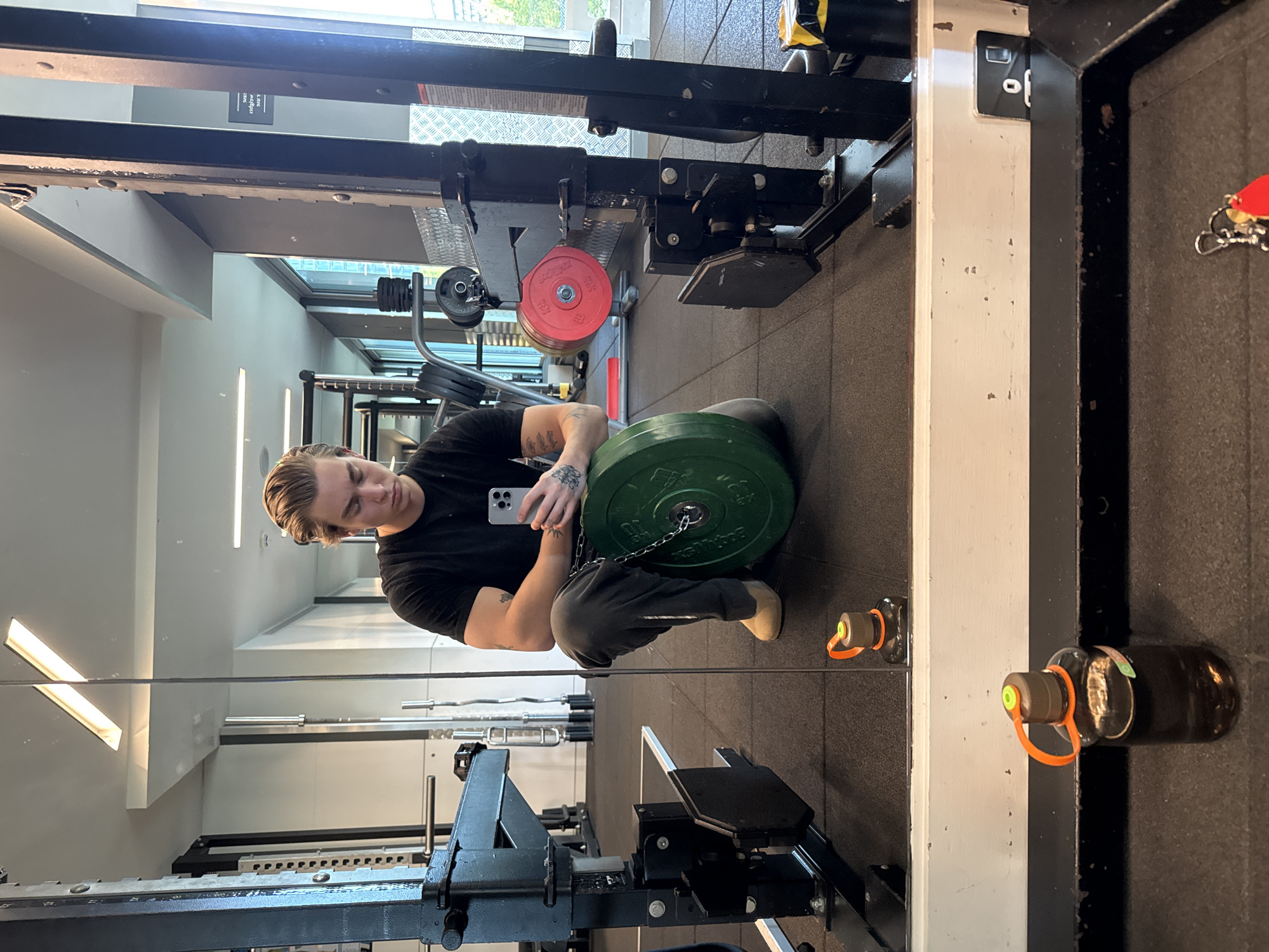 This full-shot mirror image captures a young male individual in an indoor gym setting, likely in London, United Kingdom. The man, with light-colored hair and wearing a black t-shirt and grey sweatpants, is shown in a squatting position. He is holding a light grey smartphone in his left hand, appearing to take a mirror selfie. A heavy green weight plate, marked "20 KG" and "Bumper," rests on the floor near him, connected by a chain that seems to be part of a weight belt he is wearing.
The gym environment features dark rubber flooring, light grey walls, and large windows that reveal an urban exterior and daylight. Various gym equipment is visible, including a squat rack or power cage, barbells, other weight plates (one red, marked "5 KG BUMPER"), and additional fitness machines. Artificial strip lighting illuminates the space.
Notable details include a dark water bottle with an orange loop and green cap on the floor to the right, and keychains with a red heart and other keys hanging from equipment. Partially visible text on a sign in the background includes "1. The..." and "o f." The image conveys a moment of activity within a modern fitness facility.