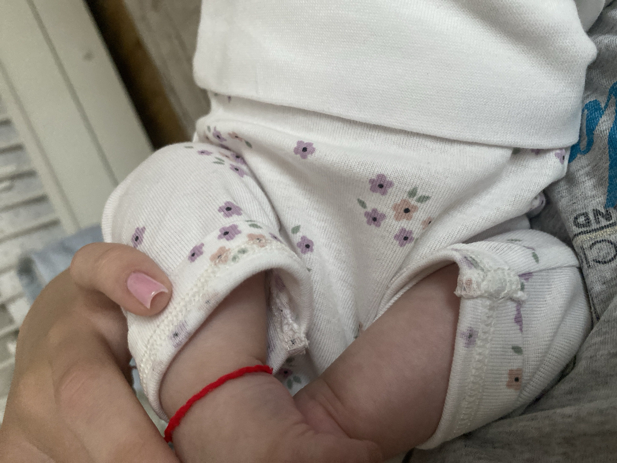 A close-up image captures a tender moment between an adult and a baby in an indoor setting, identified as Blagoevgrad, Bulgaria. The central subjects are a baby's lower legs and feet, held gently by an adult's hand.
The baby is dressed in a soft, white sleepsuit or pajama, adorned with a delicate pattern of small, scattered flowers in shades of purple and peach/pink, accented by tiny green leaves. The fabric appears soft and comfortable, with knitted cuffs folded back at the ankles. A thin, bright red string is tied around one of the baby's ankles, clearly visible beneath the adult's hand.
The adult's hand, positioned to support or caress the baby's foot, features a light pink painted fingernail on the thumb. The skin tones of both the baby and the adult suggest a light complexion.
The background is softly blurred, hinting at a domestic environment. Elements like light-colored furniture or walls, and what appears to be a hint of wood grain, are vaguely discernible to the left. To the right, another piece of fabric, possibly a grey blanket or clothing item, is visible, featuring a faint, blurry blue text pattern. While not entirely clear, a closer inspection in the crops reveals letters such as "GN" on this grey material.
The overall scene evokes feelings of warmth, care, and affection, highlighting the bond between the adult and the infant. The activity depicted is one of gentle holding or comforting. No specific time of day or weather conditions are evident from the indoor scene.