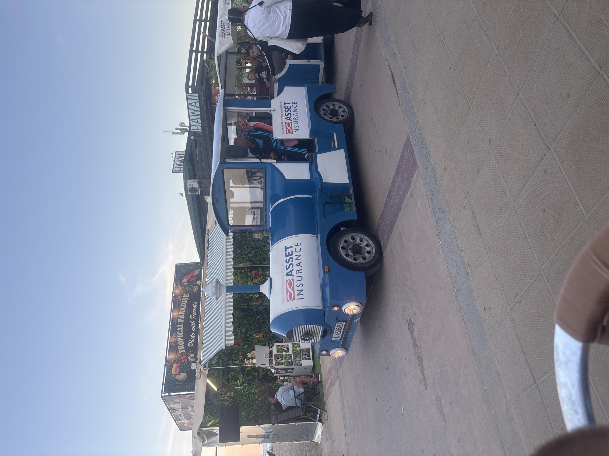In Nesebar, Bulgaria, a blue and white tourist train is parked on a paved walkway near a beach. The train has "ASSET INSURANCE" printed on its side. Behind the train, there is a canopy with a sign that reads "TROPICAL PARADISE Photo with Parrots." Further back, a building has "HAWAII" written on it. Several people are visible: two individuals are sitting in chairs near the "Tropical Paradise" stand, and others are on the train and walking nearby. The sky is clear and blue, indicating daylight. The scene suggests a tourist attraction or entertainment area.