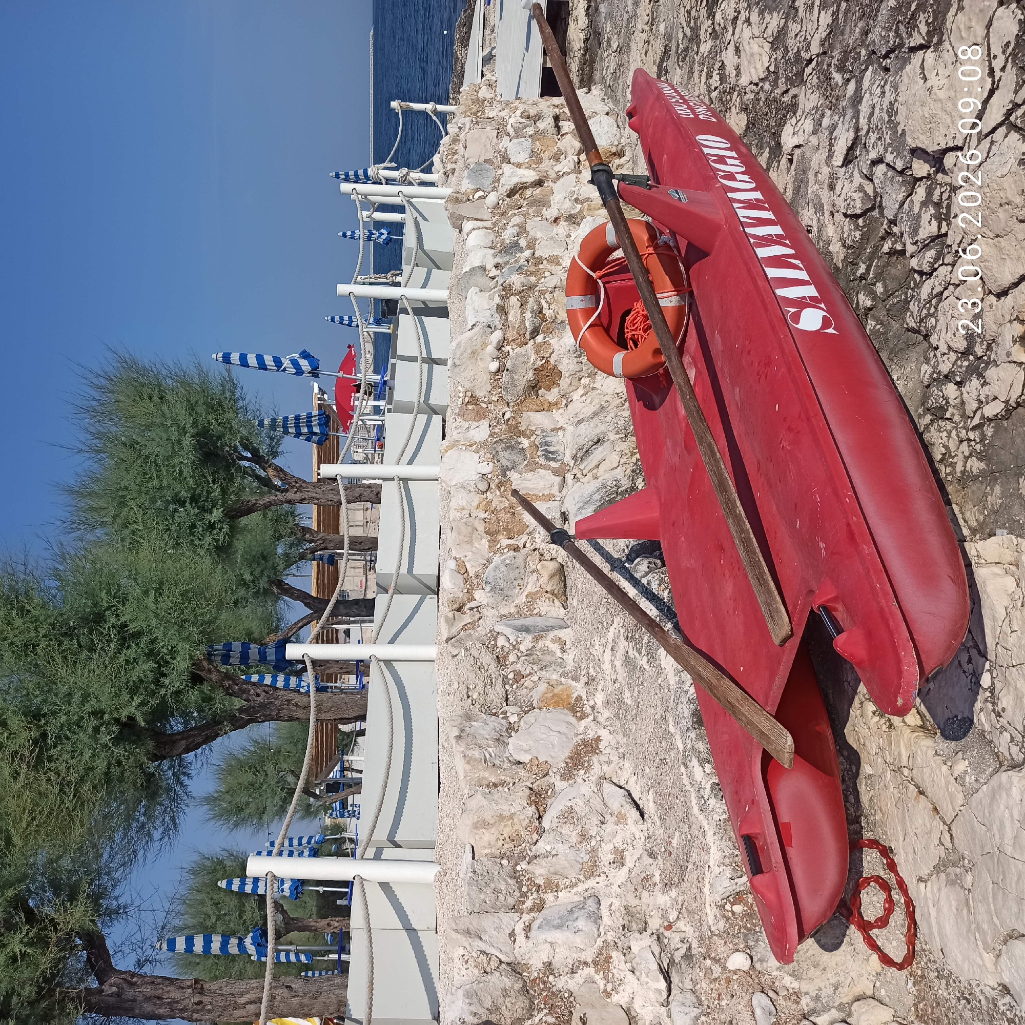 The image captures a bright, sunny day at a beach in Molfetta, Italy. In the foreground, a red, double-hulled inflatable boat, labeled "SALVATAGGIO" (meaning "Rescue" in Italian), rests on a rocky shore. Two wooden oars are laid across the boat, and an orange life preserver with a rope is visible. The boat appears to be a rescue vessel, possibly for a beach concession.
Behind the boat, a low wall constructed of rough, light-colored stones separates the rocky beach from a raised area. This raised area features a white railing with rope accents, separating a walkway or lounge area from the beach. Several closed beach umbrellas with blue and white stripes are arranged behind the railing, along with some blue beach chairs and a small wooden structure, possibly a lifeguard station or kiosk. A bright red umbrella is also visible further down the beach.
In the background, the blue expanse of the Adriatic Sea stretches to the horizon, with a distant breakwater or pier visible. Lush green trees with fine foliage, likely tamarisks, line the area behind the beach infrastructure. The sky is a clear, deep blue. The overall scene depicts a well-maintained beach facility on a calm day. A timestamp in the lower right corner reads "23.06.2026 09:08."
