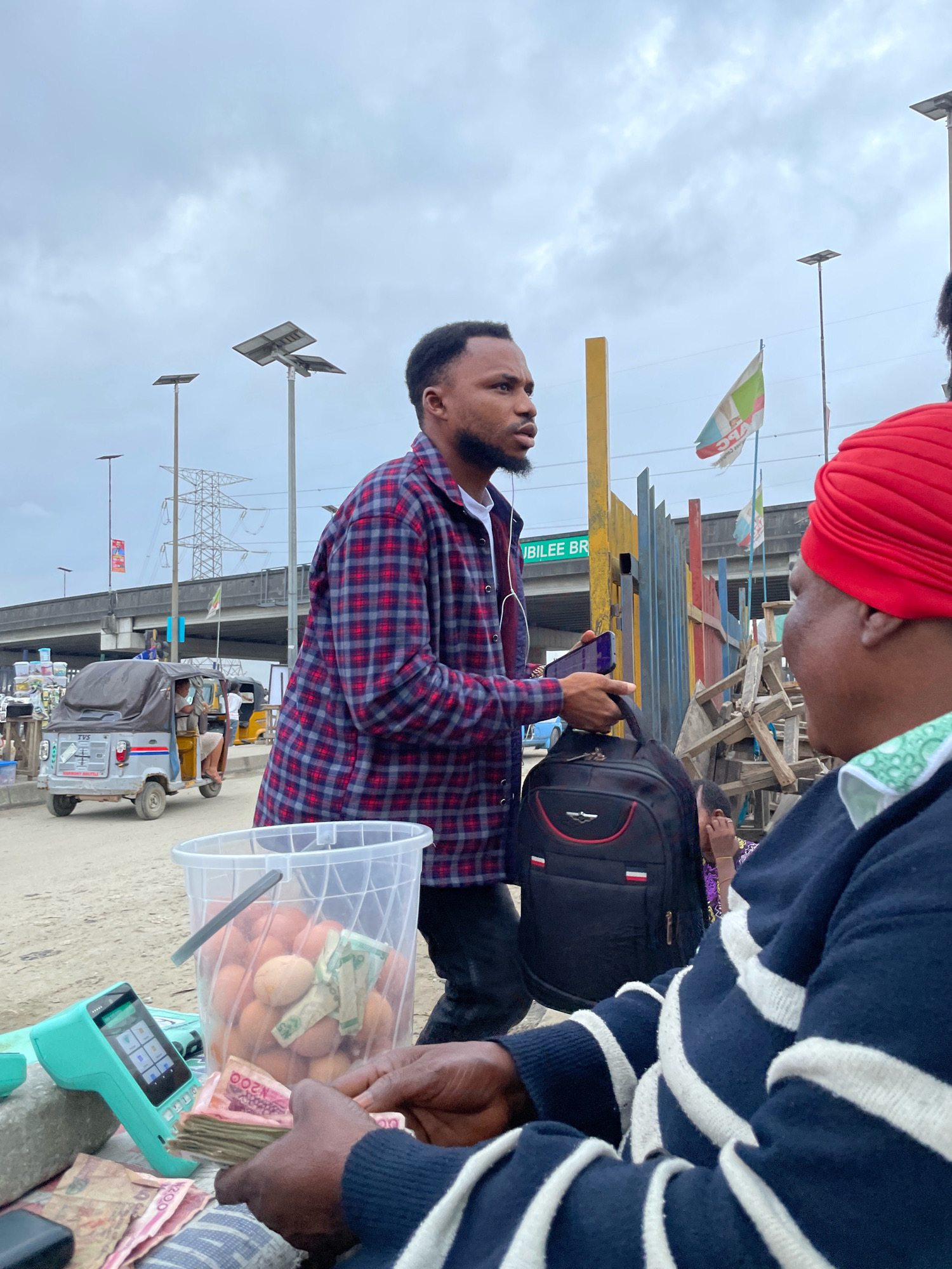 Two individuals are engaged in a transaction outdoors in Ajah, Nigeria. On the left, a man wearing a red and blue plaid shirt and dark pants stands holding a smartphone and a black backpack. He has headphones in his ears. On the right, another person, facing away from the camera and wearing a dark blue and white striped sweater and a red head covering, is counting a stack of Nigerian Naira currency. In front of them, a teal-colored card reader sits on a gray surface next to a clear plastic bucket filled with eggs and more Naira bills. The background features a street scene with a tricycle, a highway overpass with "JUBILEE BR" signage, utility poles with solar panels, and colorful metal fencing. The sky is overcast.