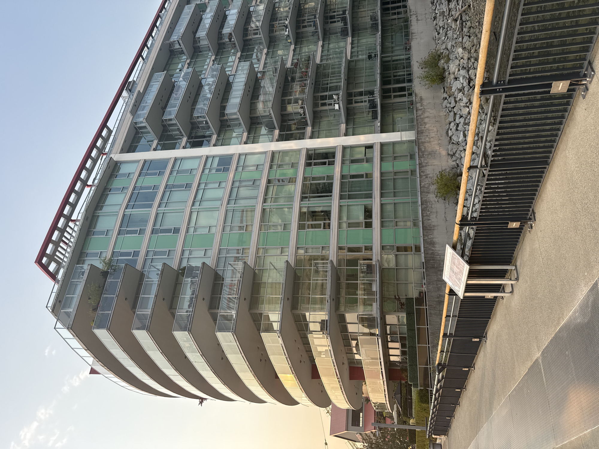 The image shows a modern, multi-story residential building with a curved facade and numerous balconies. The building has a white exterior with accents of green-tinted windows. A red, angular roof structure is visible at the top of the building. In the foreground, there is a paved walkway with a black metal railing. To the right of the walkway, there is a concrete retaining wall and a rocky embankment. The scene appears to be a waterfront area. The sky is a pale, hazy blue, suggesting it might be late afternoon or early morning. In the distance, to the left, a smaller building with a red roof is visible. There are no people visible in the image. The location is identified as the Westview Marina, North Vancouver, Canada.