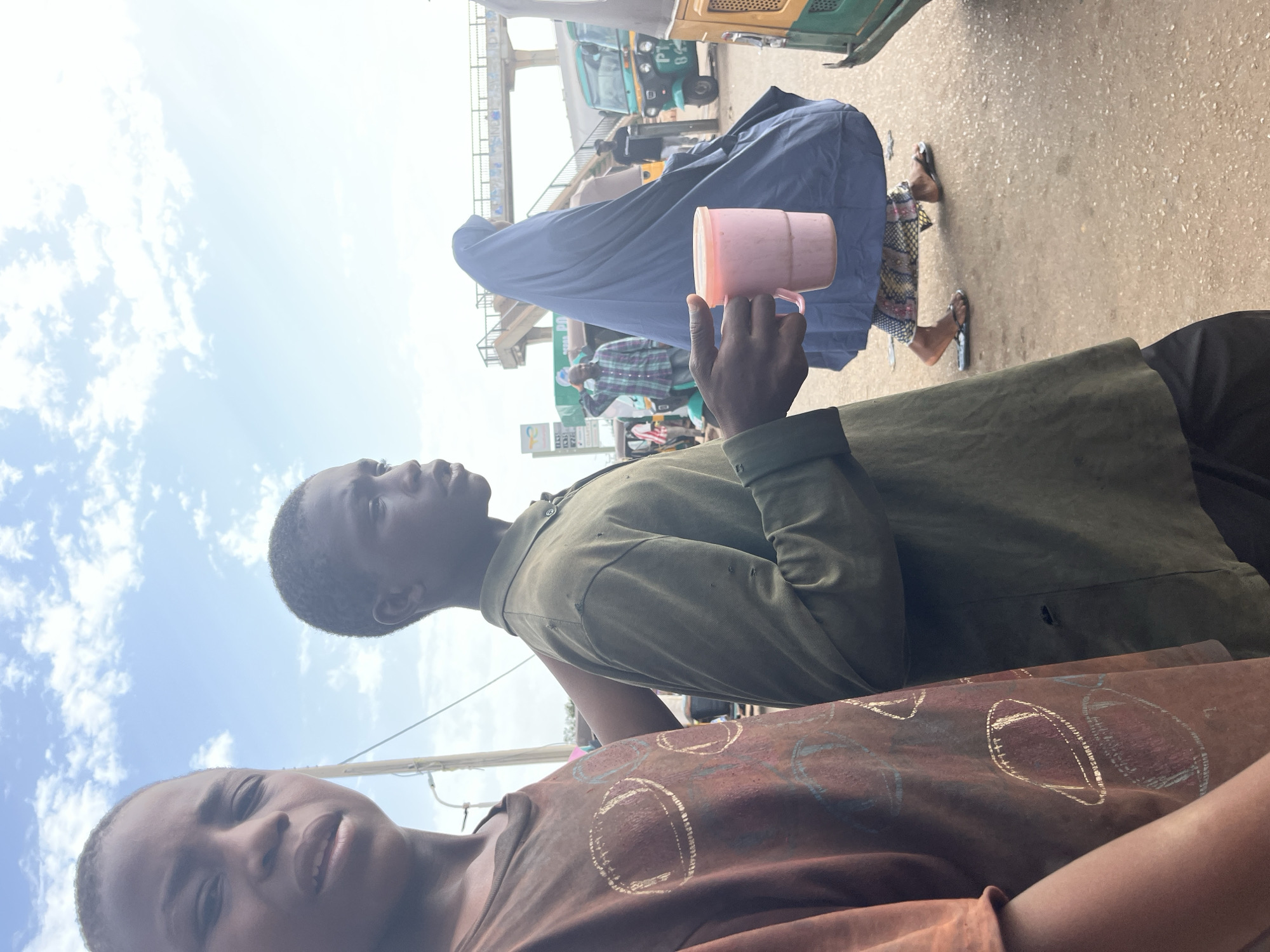 In Gura Top, Nigeria, two young boys are pictured walking side-by-side. The boy on the left wears a brown t-shirt with a repeating football design. The boy on the right is wearing a green shirt and is holding a pink cup with a pink liquid. Behind them, a woman in a blue hijab walks past, followed by several auto-rickshaws and a pedestrian bridge. The scene takes place on a dirt road under a partly cloudy sky.