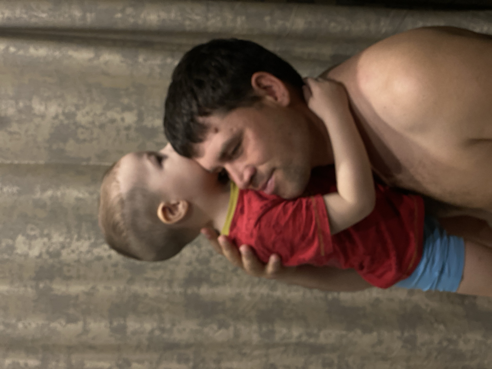 An adult male with dark hair and a light complexion, shirtless, holds a young child with light brown hair. The child, wearing a red short-sleeved shirt with a yellow trim and light blue shorts, is embracing the adult, with their head resting against the adult's head, appearing to nuzzle or kiss the adult's temple. The adult's right arm supports the child, and his gaze is directed towards the child. The scene occurs indoors against a textured, light brown or beige background, possibly wallpaper or a curtain. The lighting is soft and ambient. The precise location within Sochi, Russia, is not identifiable from the visual elements presented in the image.