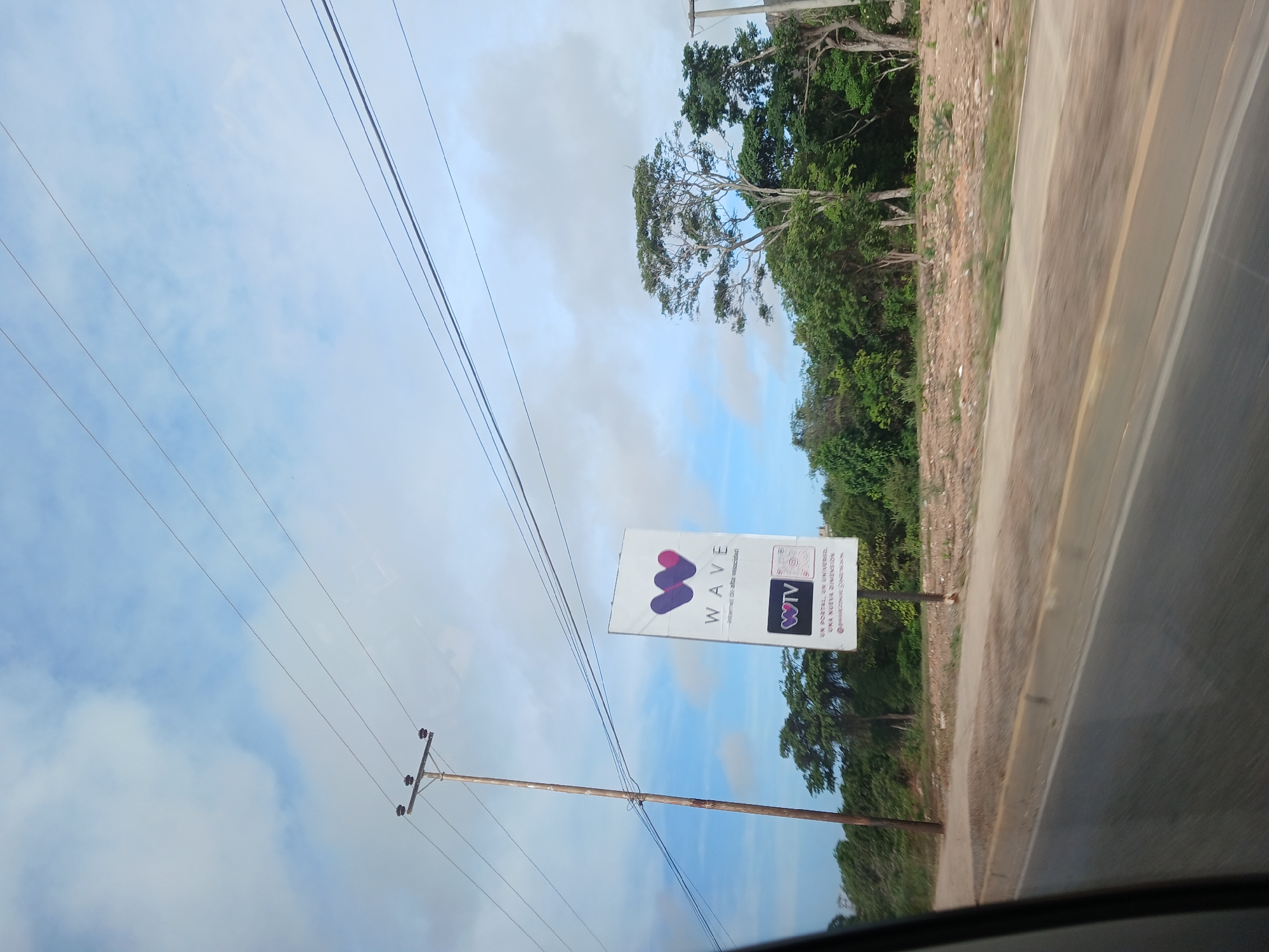 This image captures a roadside view in El Valle del Espíritu Santo, Venezuela. A wooden utility pole stands on the left, with power lines stretching across the sky. To the right of the pole is a tall, white billboard advertising "WAVE" internet service. The billboard features the WAVE logo, the text "WAVE - Internet de alta velocidad," and smaller logos for "WTV" and a QR code. Below this, more text reads "UN PORTAL, UN UNIVERSO, UNA NUEVA DIMENSION" followed by a web address and social media handle. The background consists of lush green trees and vegetation, with a strip of bare, sandy ground between the roadside and the foliage. The road itself is paved with a curb. The sky is partly cloudy, with blue sky visible between the white clouds. The perspective suggests the photo was taken from a moving vehicle.
