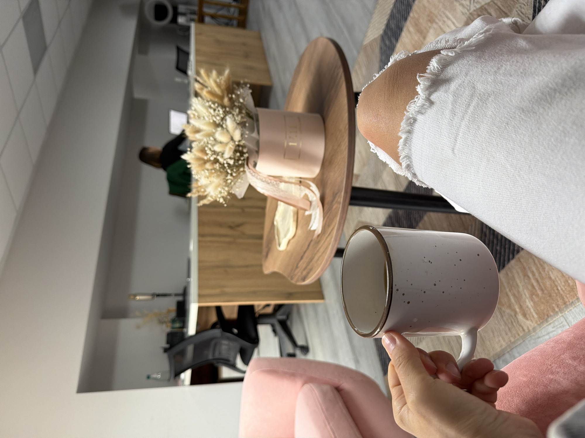 This vertically rotated image captures an indoor scene, likely a modern office or lounge area in Chaikovsky, Russia. The foreground features a person's hand holding a white ceramic mug adorned with small dark speckles and a golden rim. The person is wearing white jeans with fashionable rips and frayed edges visible near the knee. Below the hand and leg, a plush pink armchair is partially visible, suggesting the person is seated and relaxing.
To the left, a round wooden side table holds a light pink cylindrical box containing a bouquet of dried flowers. The bouquet primarily consists of delicate, feathery light brown grasses (resembling bunny tail grass) mixed with small white dried filler flowers. Gold lettering, which appears to spell "FLEUR," is visible on the side of the pink box, and a light pink ribbon is draped around it.
In the blurred background, an office setting unfolds. A person with dark hair, wearing a dark green top, is seated at a light wooden desk or counter, seemingly engaged with a computer monitor. The office features light-colored walls, possibly with subtle patterns, and a patterned rug in neutral tones of beige, brown, and grey is visible on the floor. Other office furniture, including chairs, can be seen further back. The overall ambiance is bright and modern, suggesting daytime. The scene depicts a moment of leisure within a professional or semi-professional environment, with one person relaxing and another working.